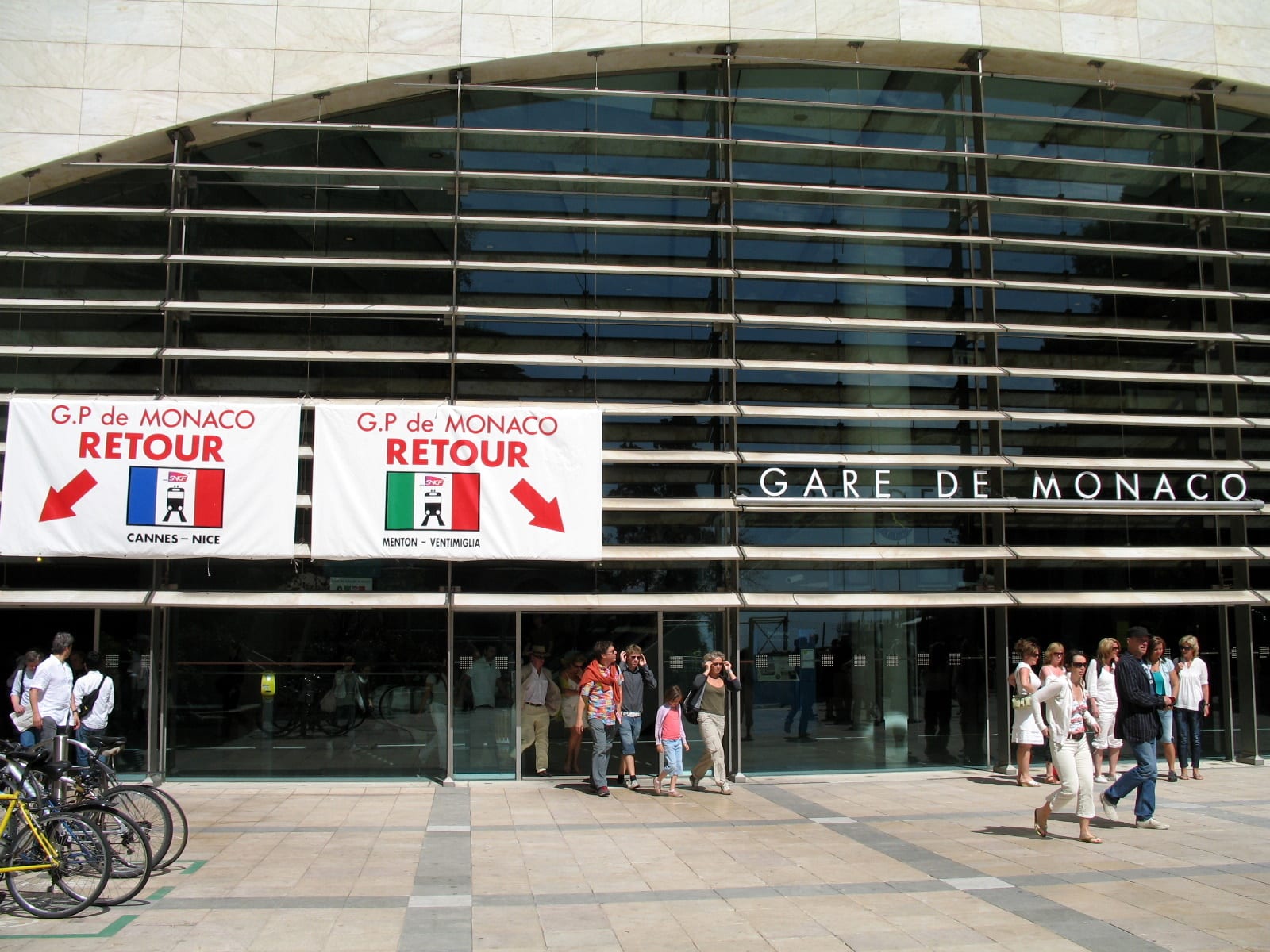 Crowds of people walking out through the glass-fronted entrance of the Gare de Monaco railway station beneath a large sign reading Gare de Monaco