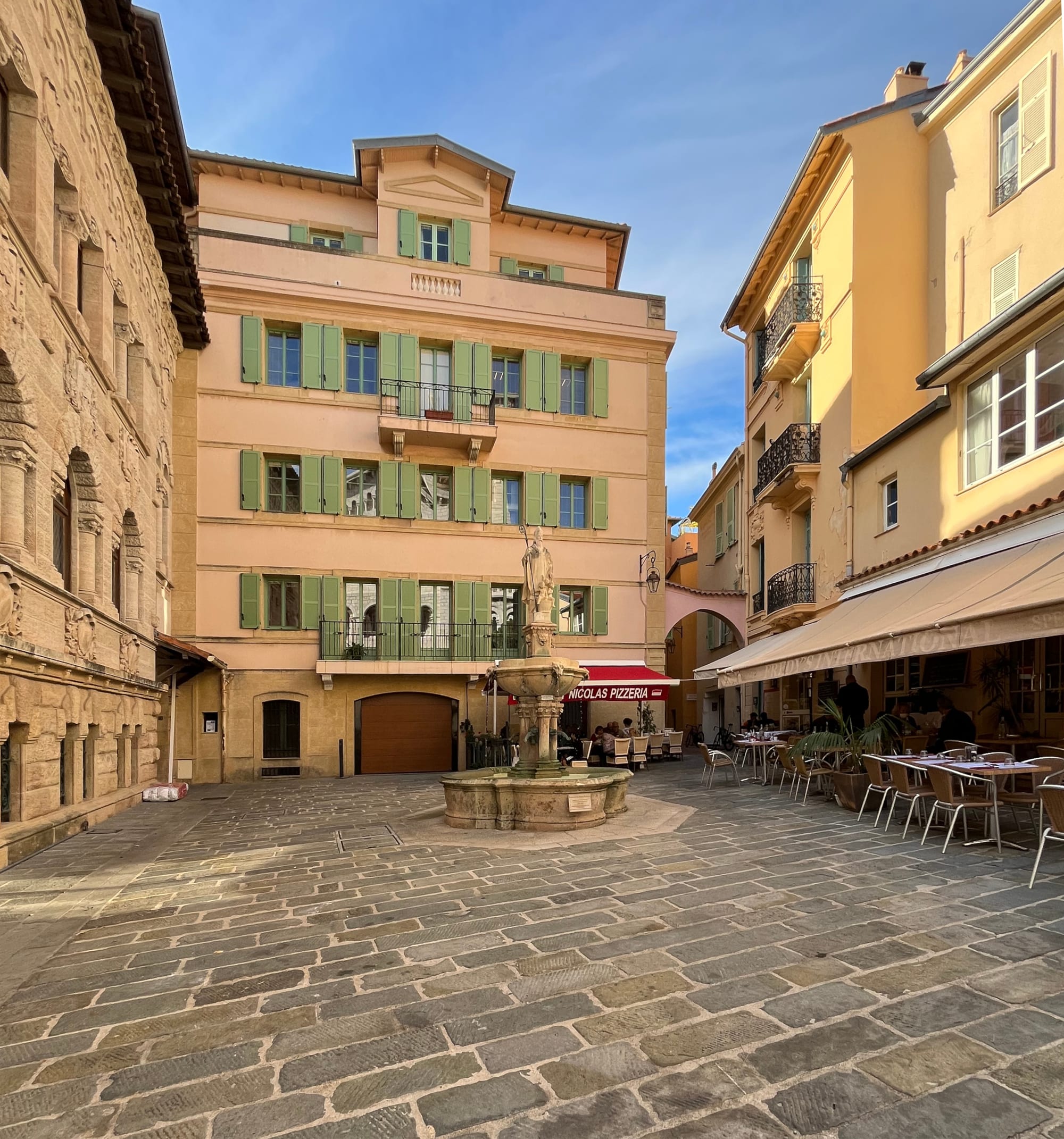 A small paved square surrounded by pastel-coloured buildings with green shutters, with a stone fountain topped by a statue in the centre and café tables set out under awnings