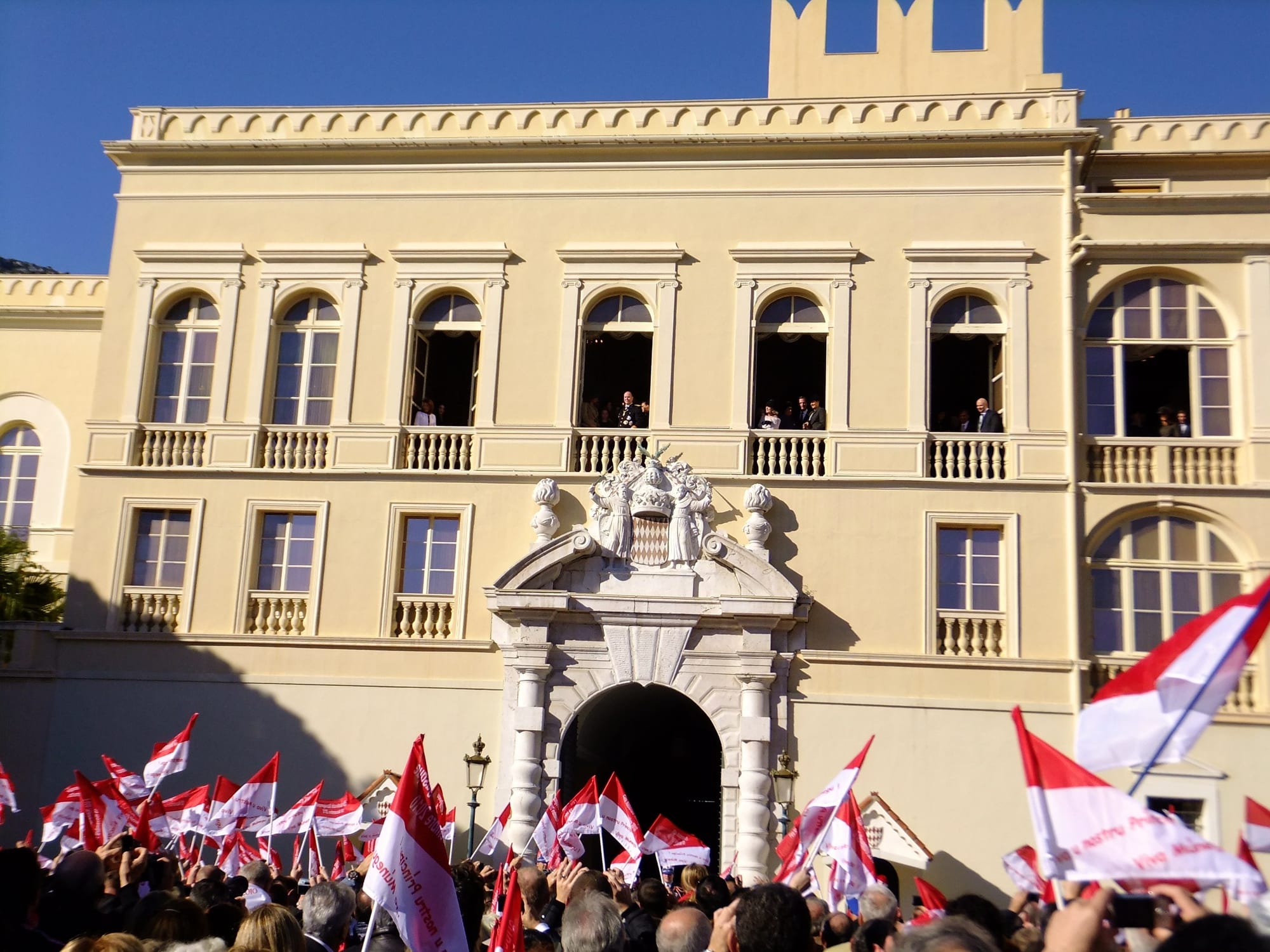 A crowd holding up many red and white flags in front of a pale yellow palace facade, with people watching from the arched windows and balconies above