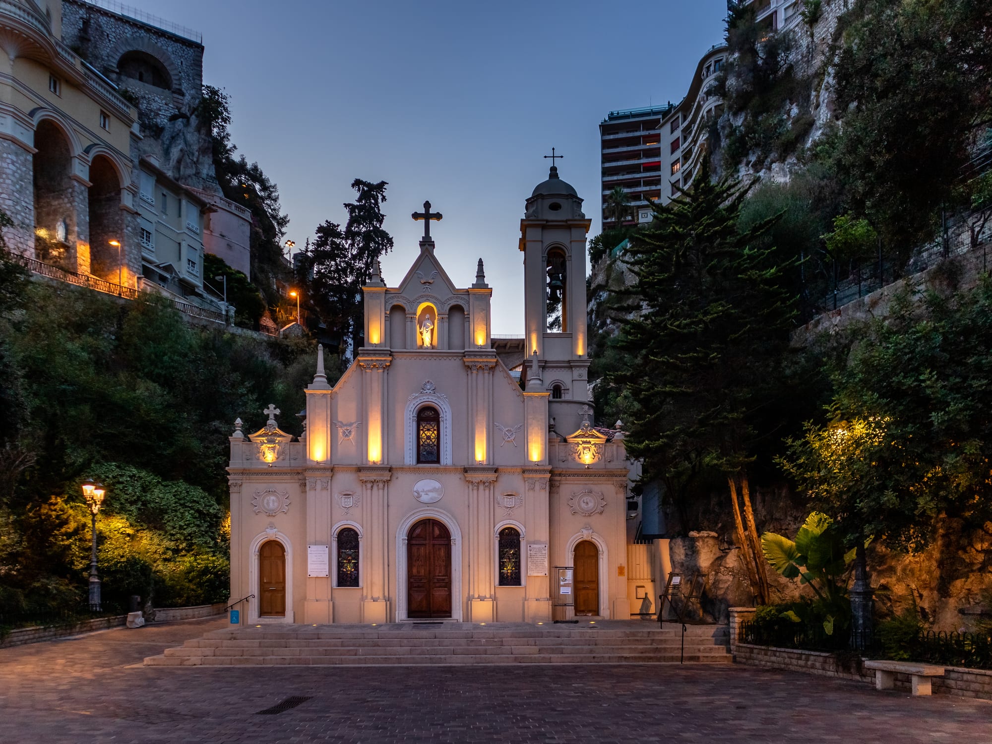 A small cream-coloured church with a bell tower and a lit facade at dusk, set against a steep, rocky and wooded hillside with apartment blocks above