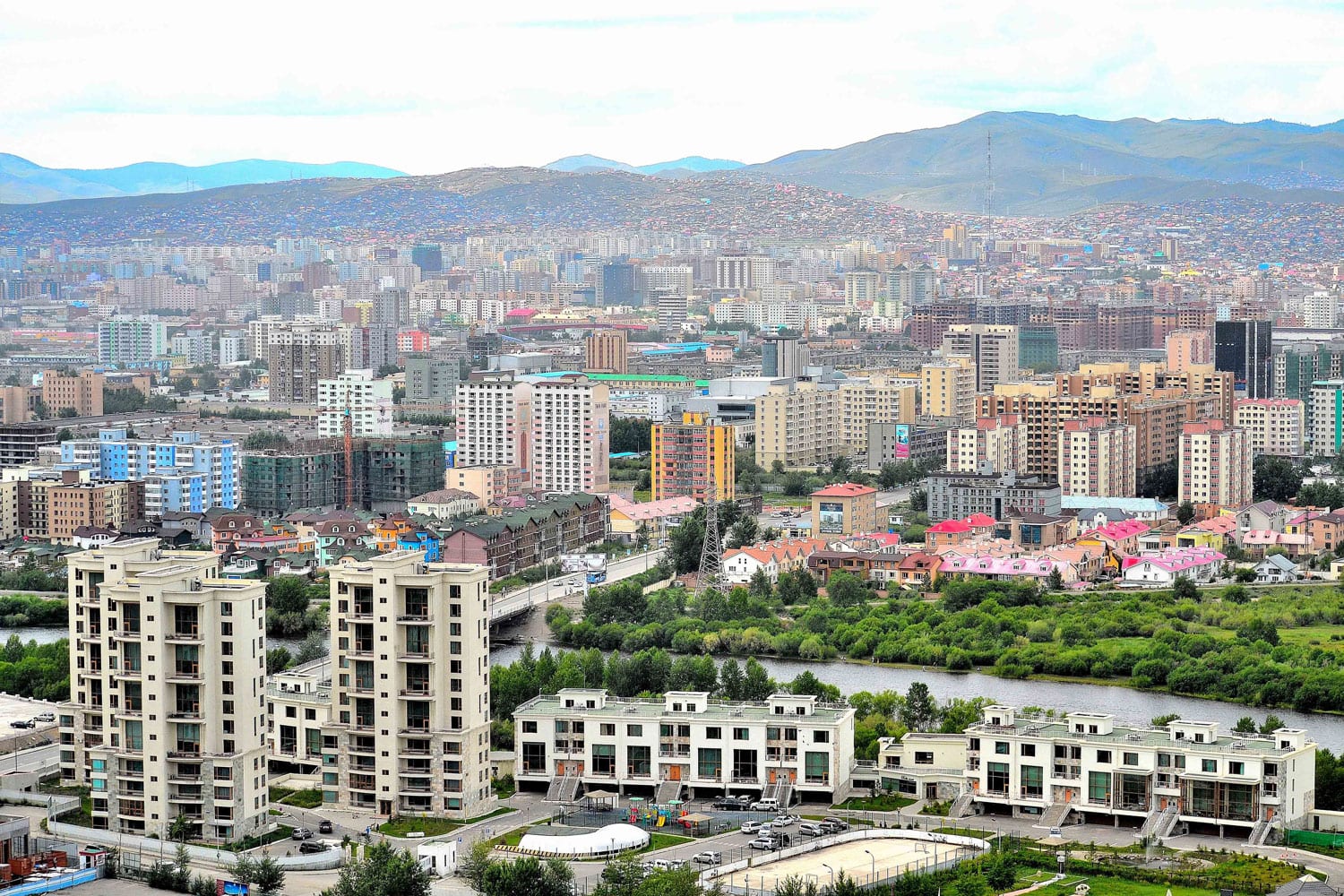 A dense skyline of high-rise apartment blocks in Ulaanbaatar spreading toward hills in the distance, with a river and lower-rise housing in the foreground