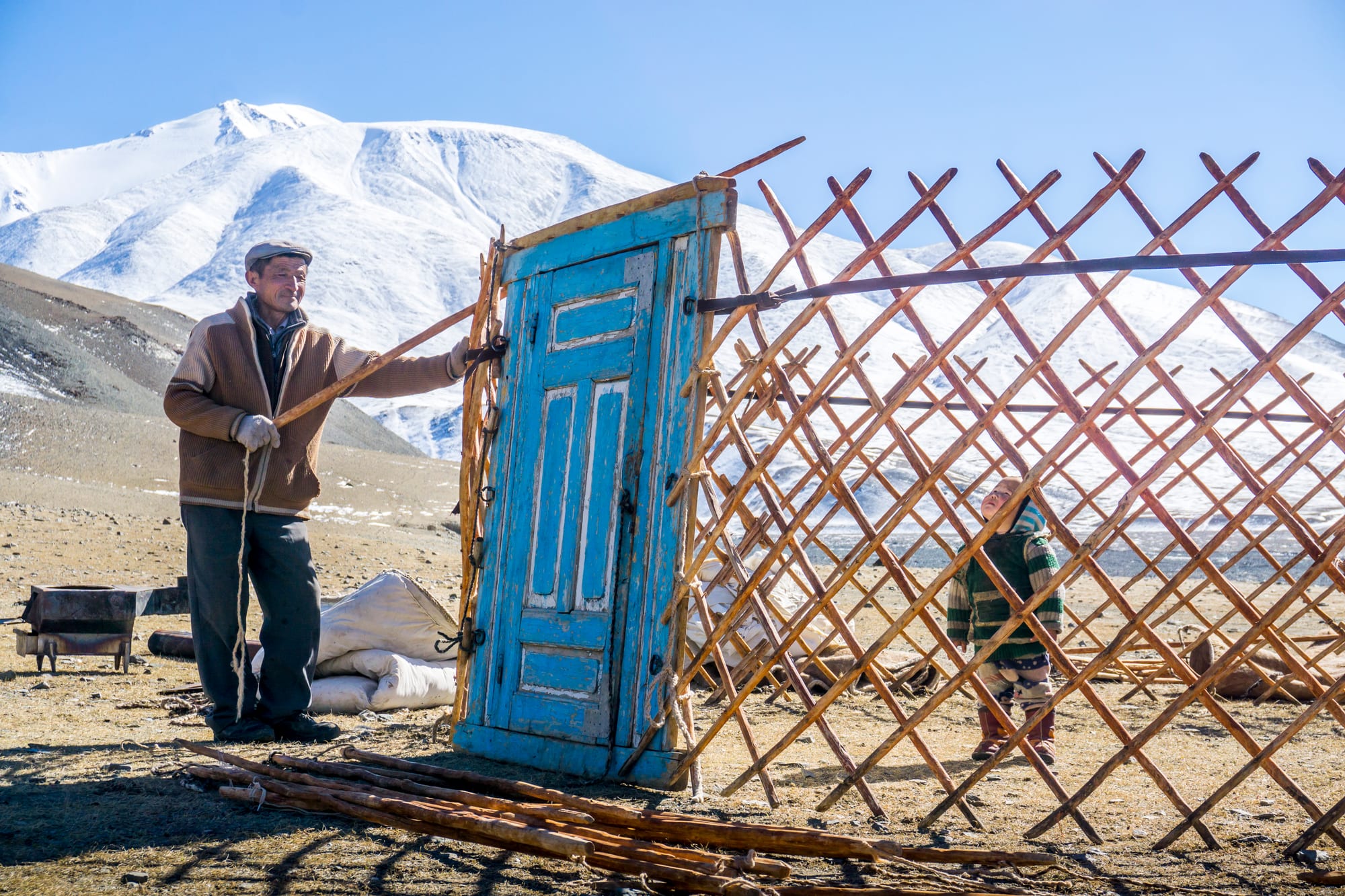 A man and a child assembling the wooden lattice frame of a ger on open ground, with snow-capped mountains behind