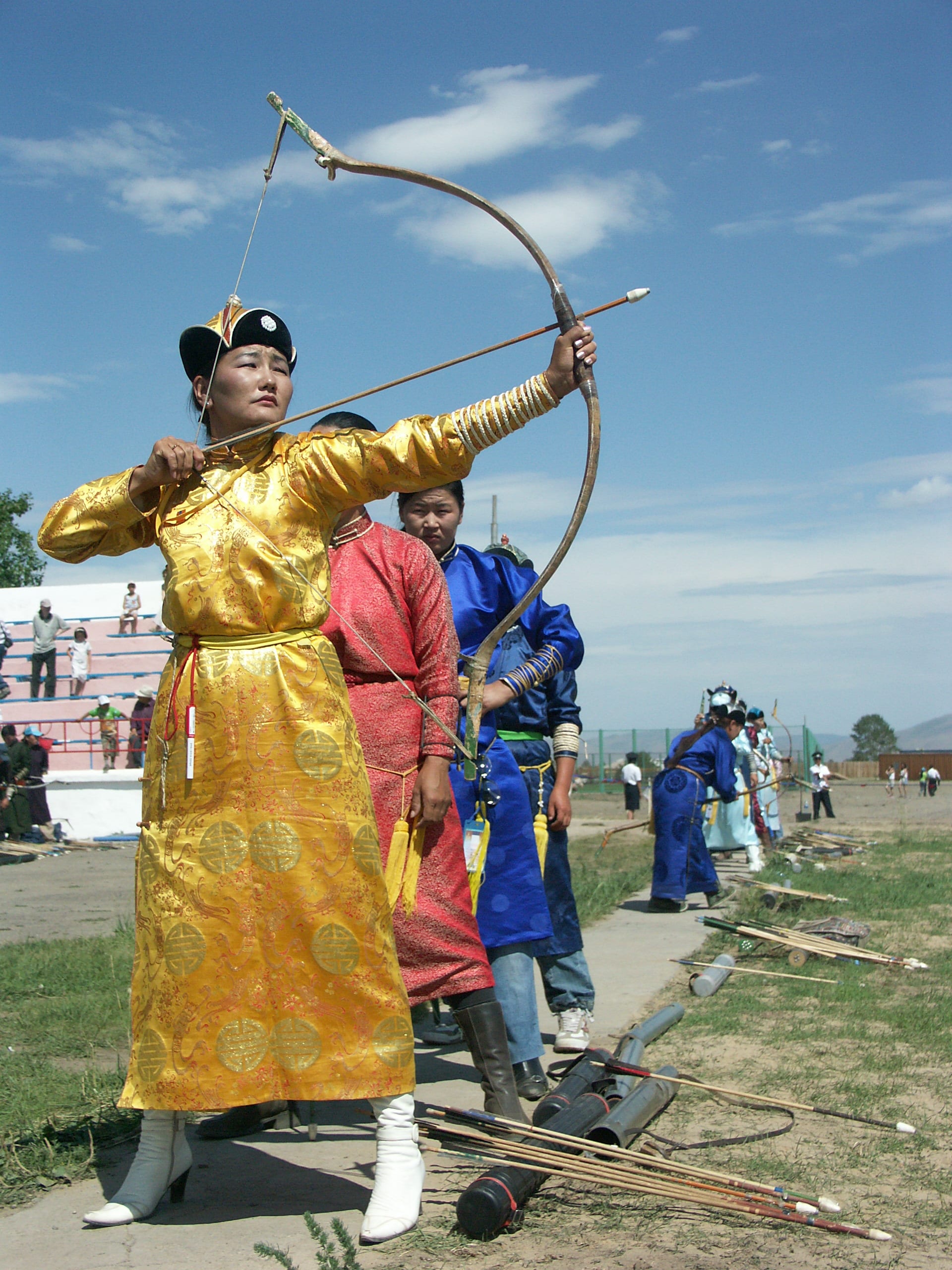 A woman in a gold brocade robe and a black and gold hat draws a long curved bow, with other archers in red and blue robes lined up behind her and arrows laid on the ground
