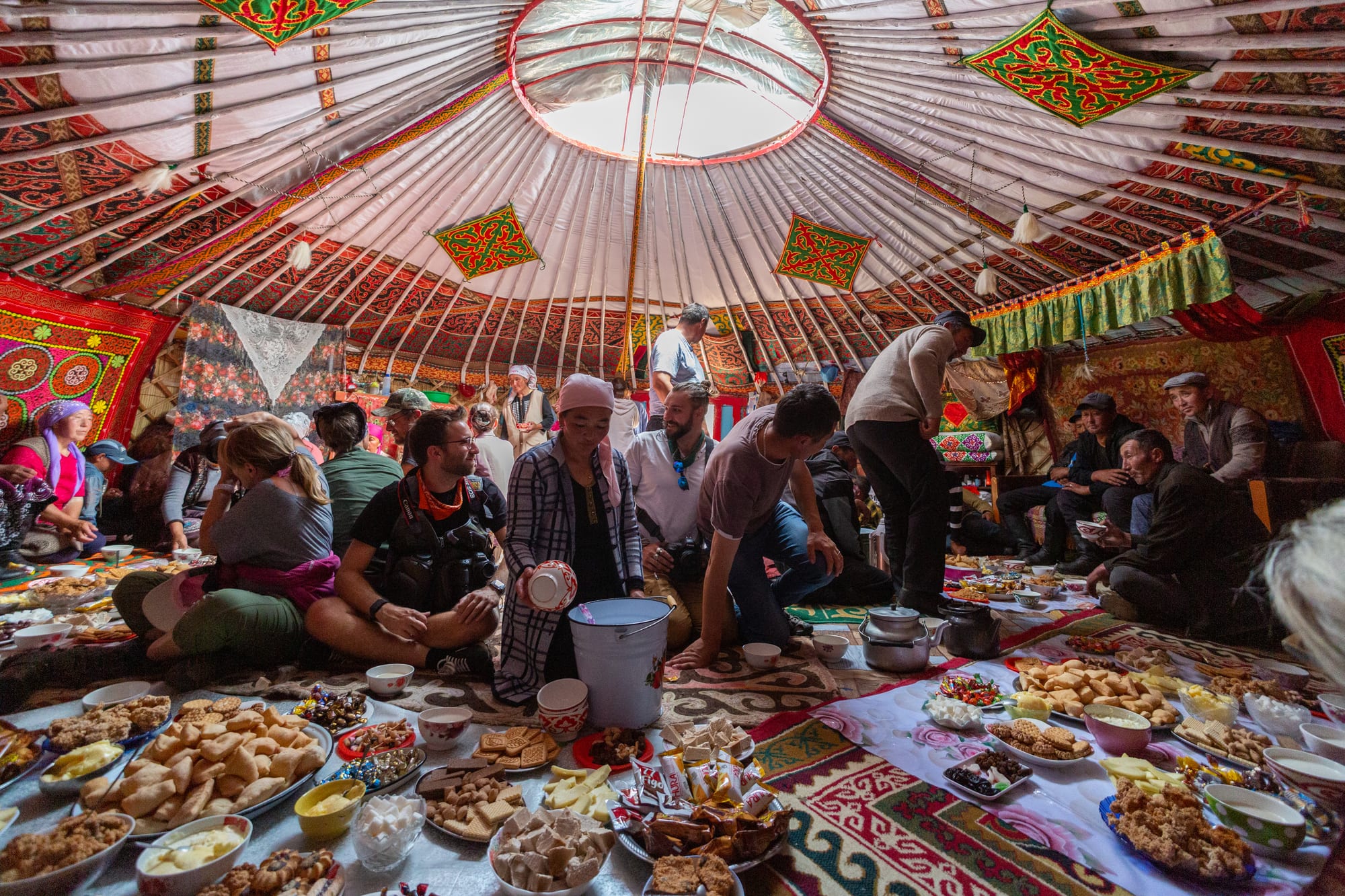 Guests sit cross-legged on patterned rugs inside a richly decorated ger around a floor spread of breads, sweets, cheeses and bowls, while a woman in a headscarf pours from a white pail