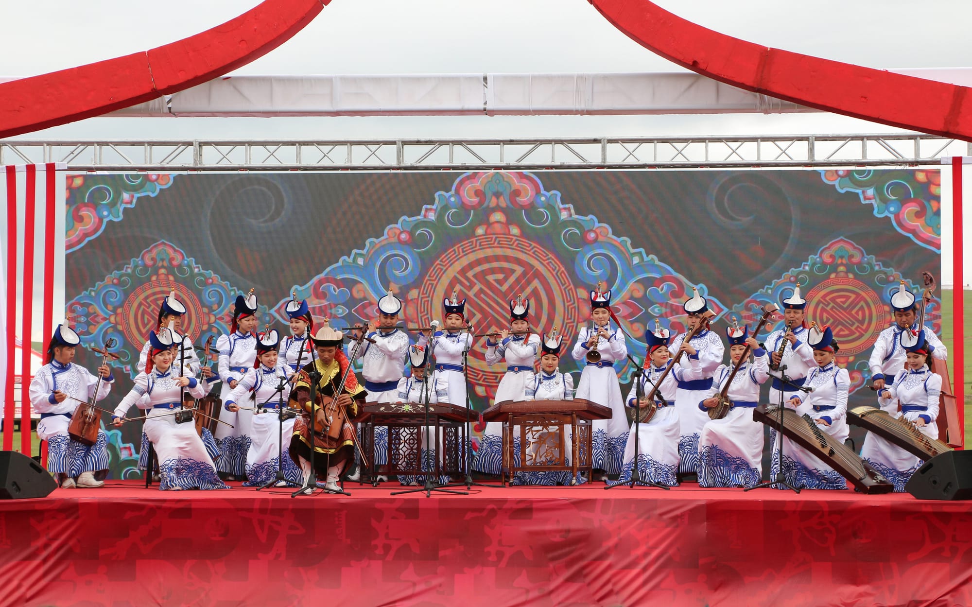 A large ensemble in white and blue robes and pointed hats plays horse-head fiddles, zithers, lutes and flutes on an outdoor stage in front of a colourful patterned backdrop