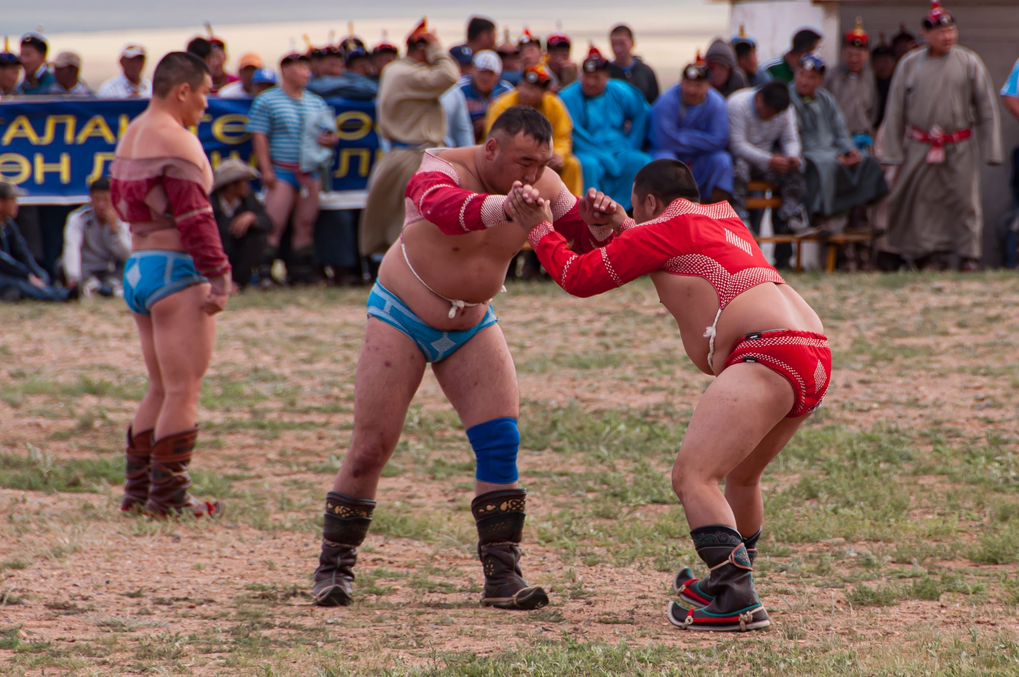 Two Mongolian wrestlers in traditional open-fronted jackets grappling at a Naadam festival