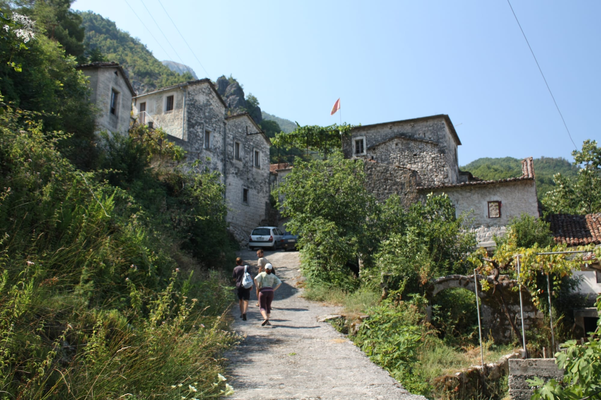 A stone-built mountain village with steep tiled roofs and a dirt lane, people walking up the path between the houses with wooded hills rising behind