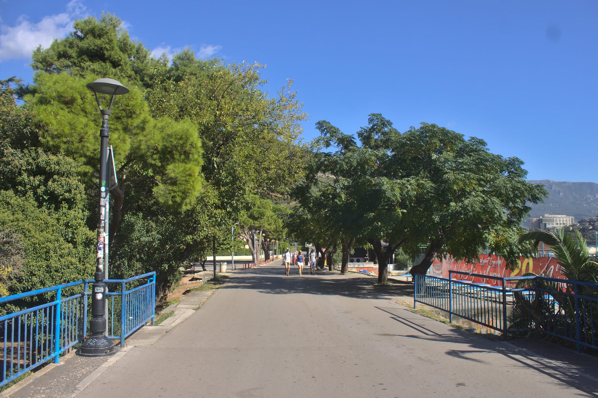 A tree-lined seaside promenade in Budva with a few people walking, blue railings, and hills in the distance