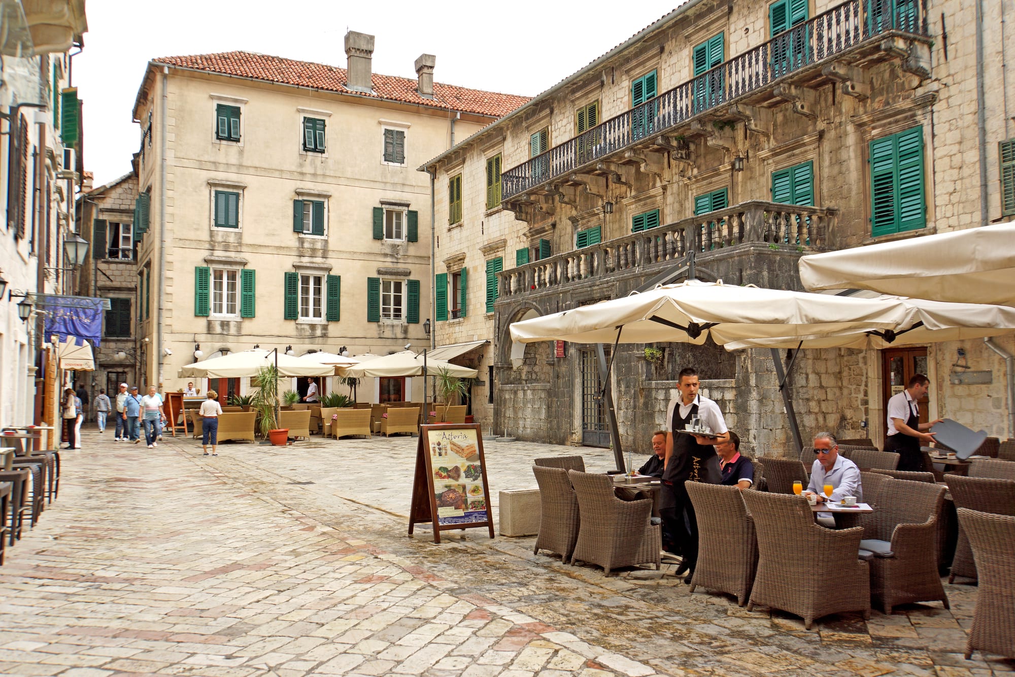 A café terrace in a stone-paved square in Kotor's old town, with waiters serving customers seated under large umbrellas