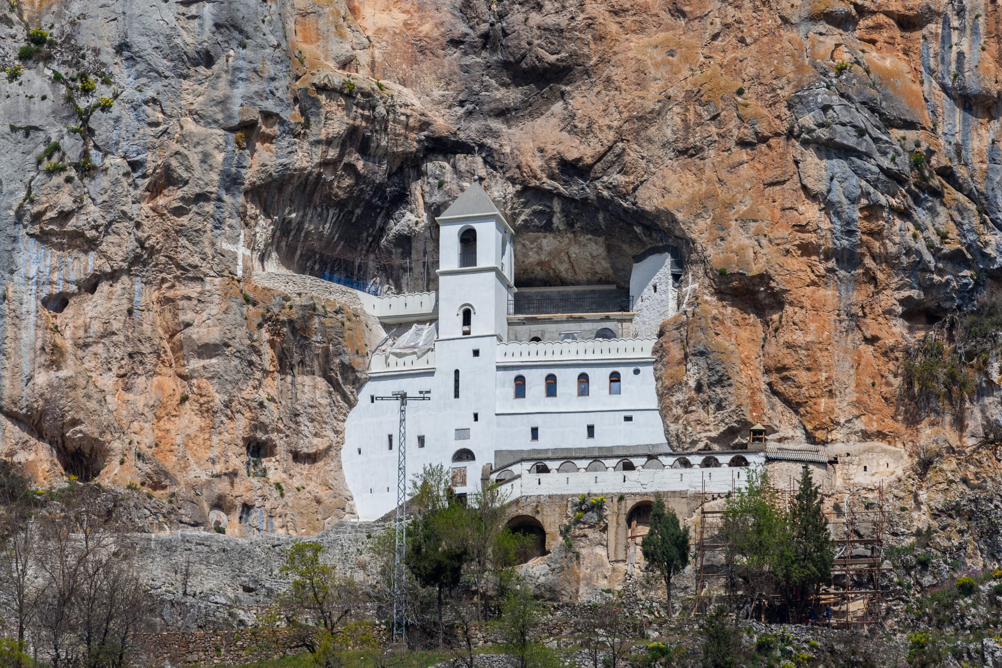 A white monastery with a small bell tower built into a hollow in a towering orange and grey cliff face, with trees and a power pylon below