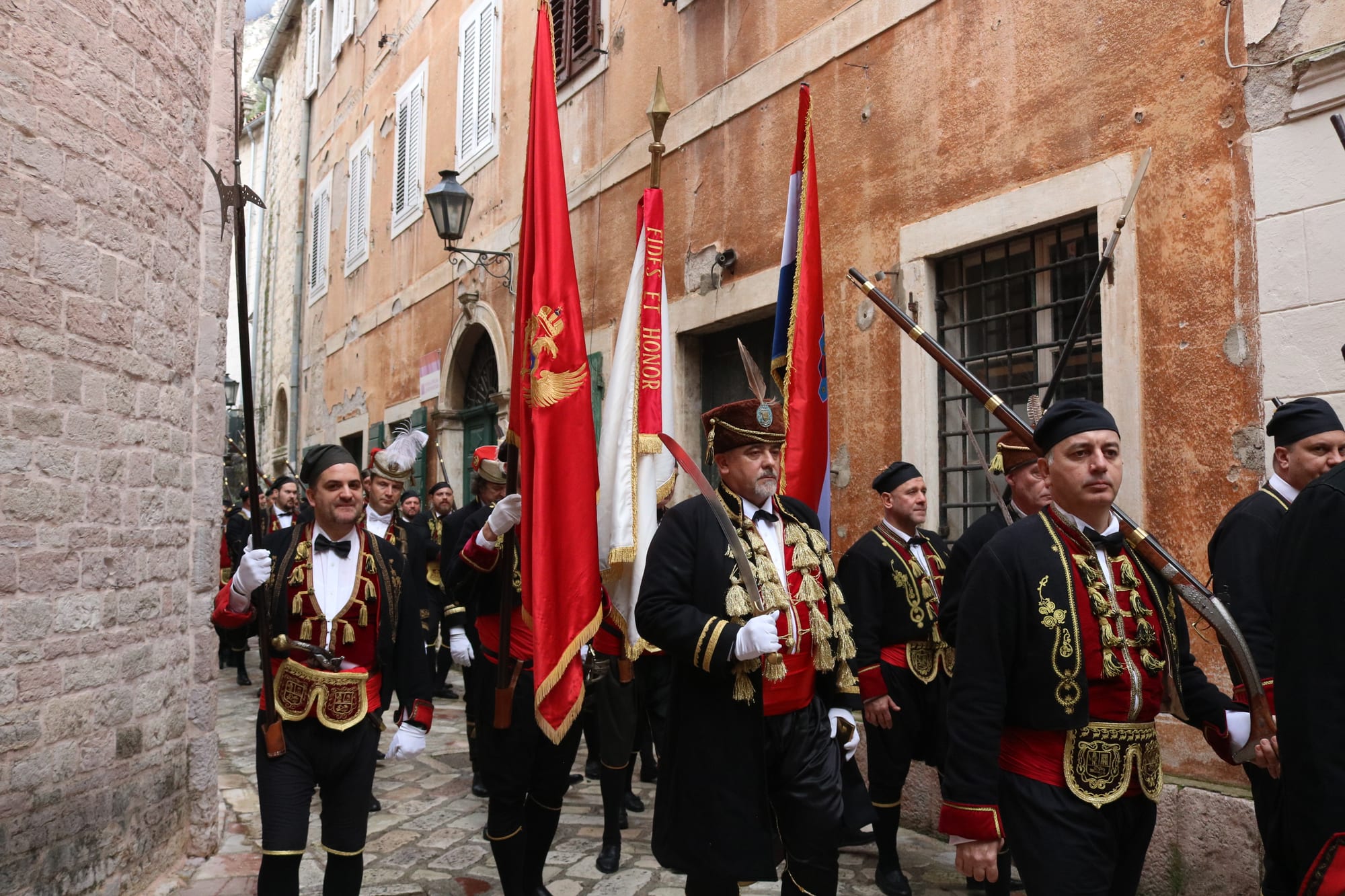 Men in black jackets with gold braid, red sashes, white gloves and black caps process along a narrow stone street carrying red flags, a white banner, halberds and old muskets