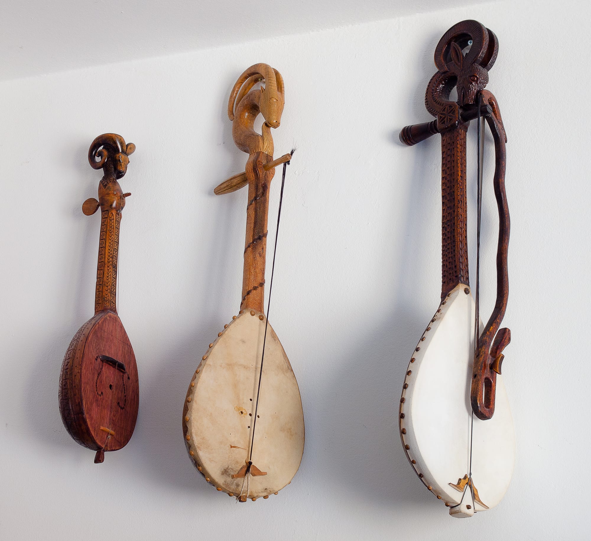 Three carved wooden stringed instruments hang on a white wall, each with a teardrop body, a single string and a neck topped with a carved animal head