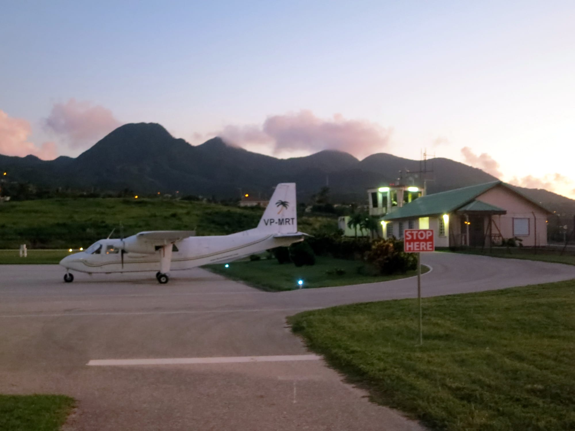 A small twin-propeller aircraft parked on the tarmac beside a low green-roofed terminal building, with volcanic hills behind