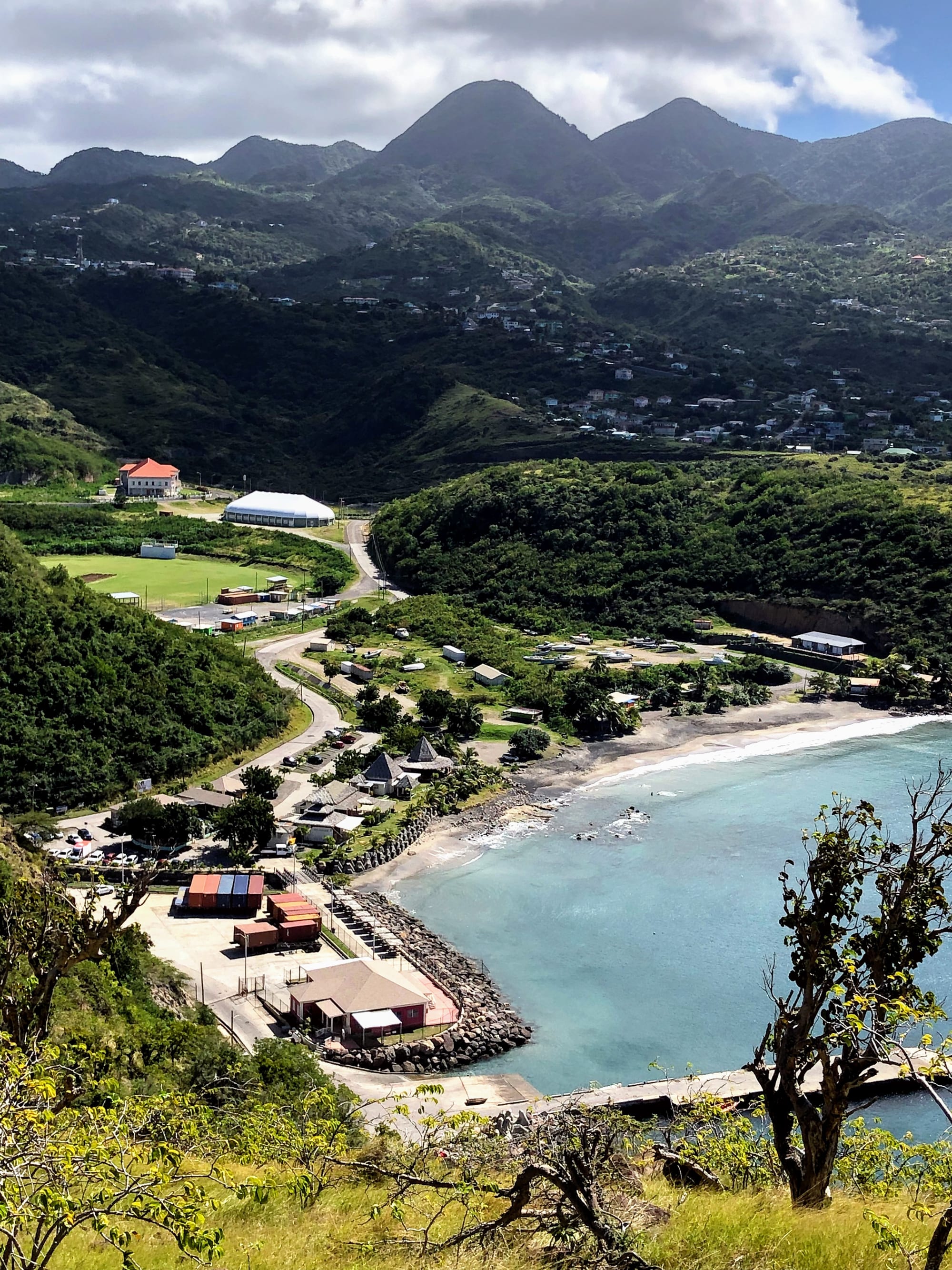 An aerial view of Little Bay, Montserrat, showing a small port, sports ground and cluster of buildings on a green bay backed by forested hills