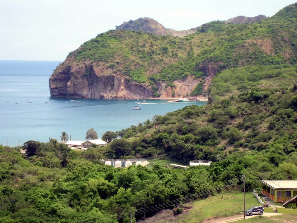 A small bay with a beach and a few low buildings, green hills behind