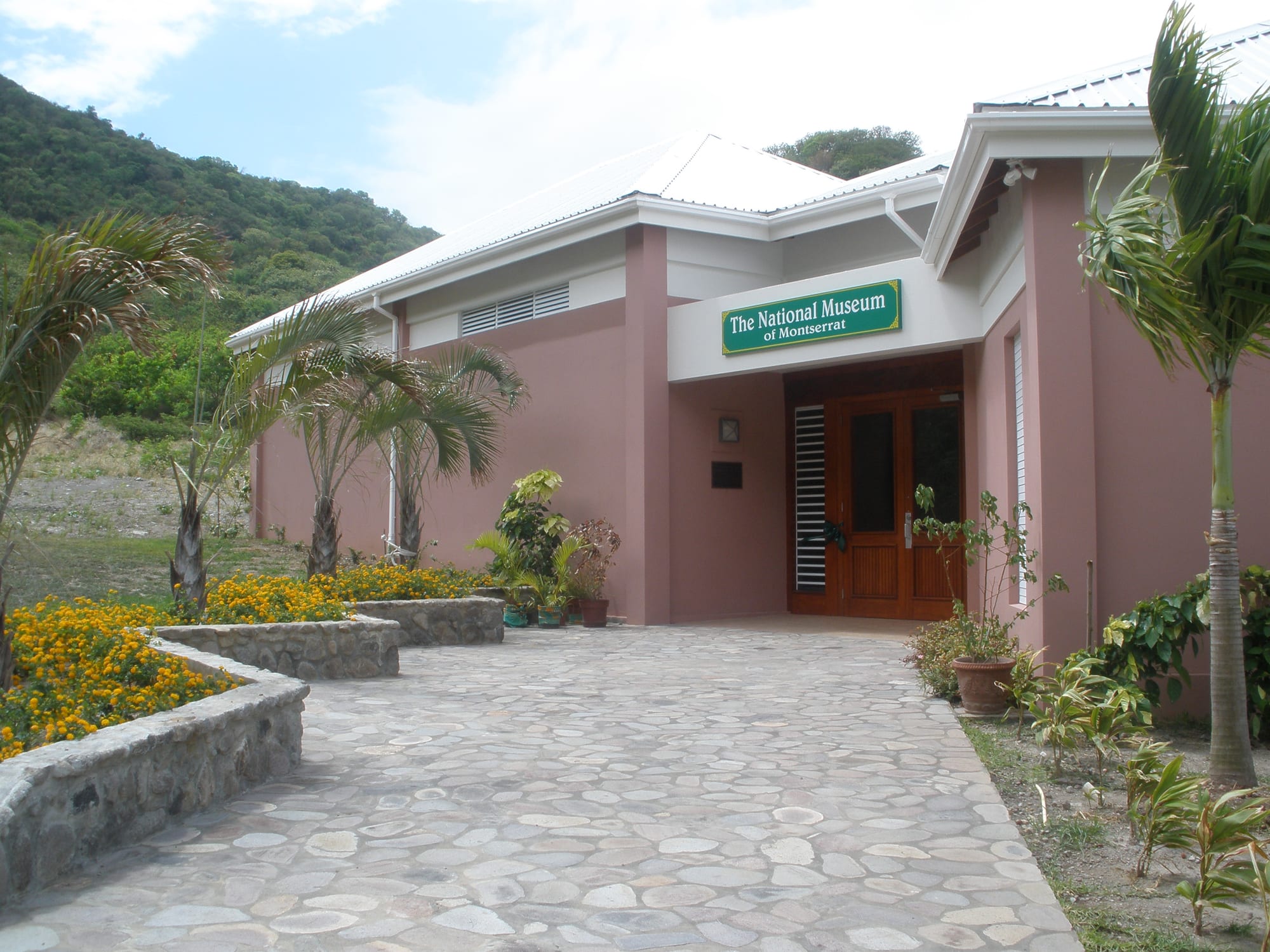 A single-storey pink building with a pale roof and a green sign reading The National Museum of Montserrat, approached by a flagstone path between flower beds and young palms