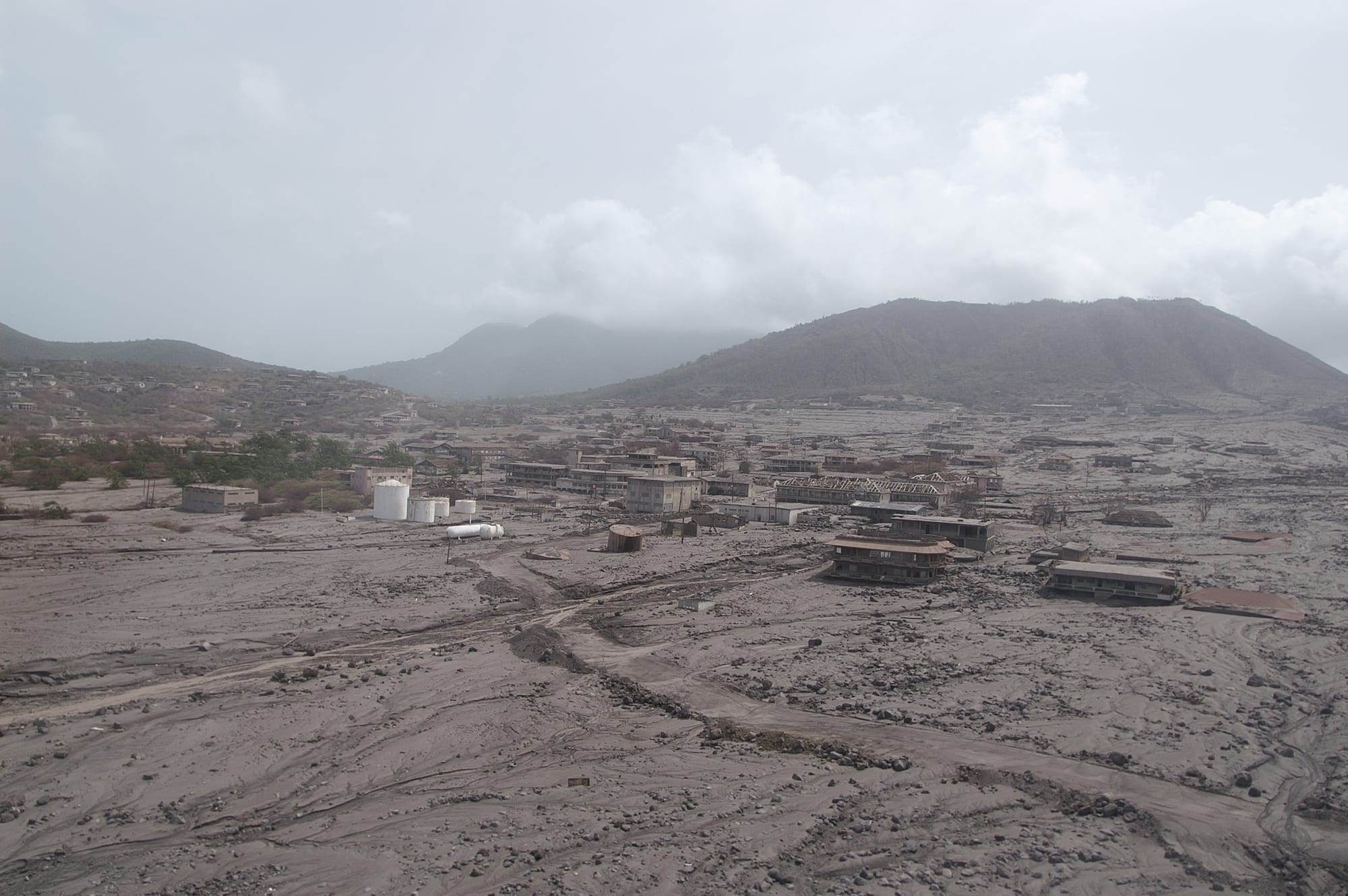 A grey plain of volcanic deposits stretches towards green hills under cloud, with the roofs and upper floors of abandoned buildings and white storage tanks half buried in the ash