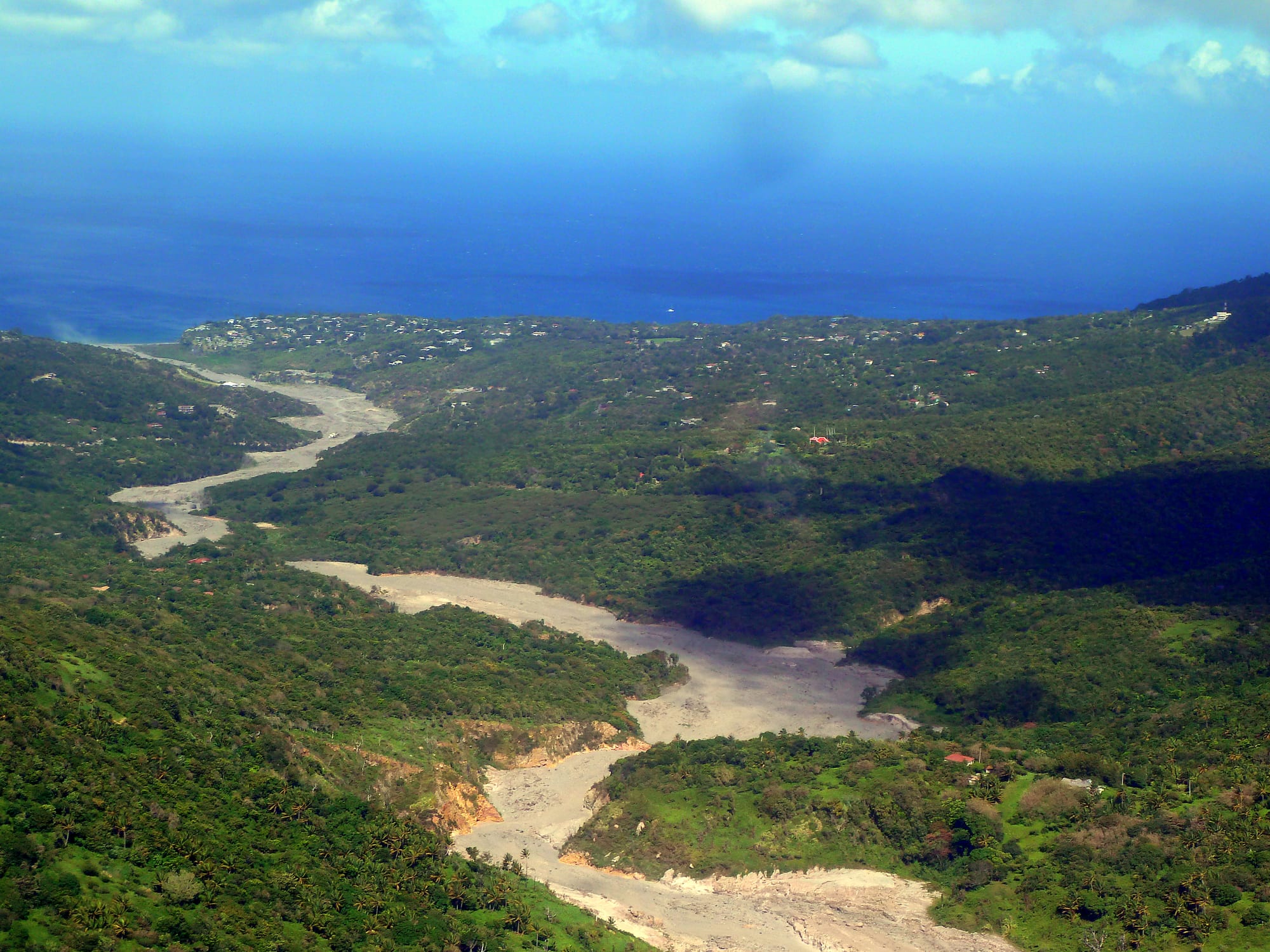 An aerial view of green wooded hills scattered with houses running down to a blue sea, with broad pale riverbeds winding through the valley