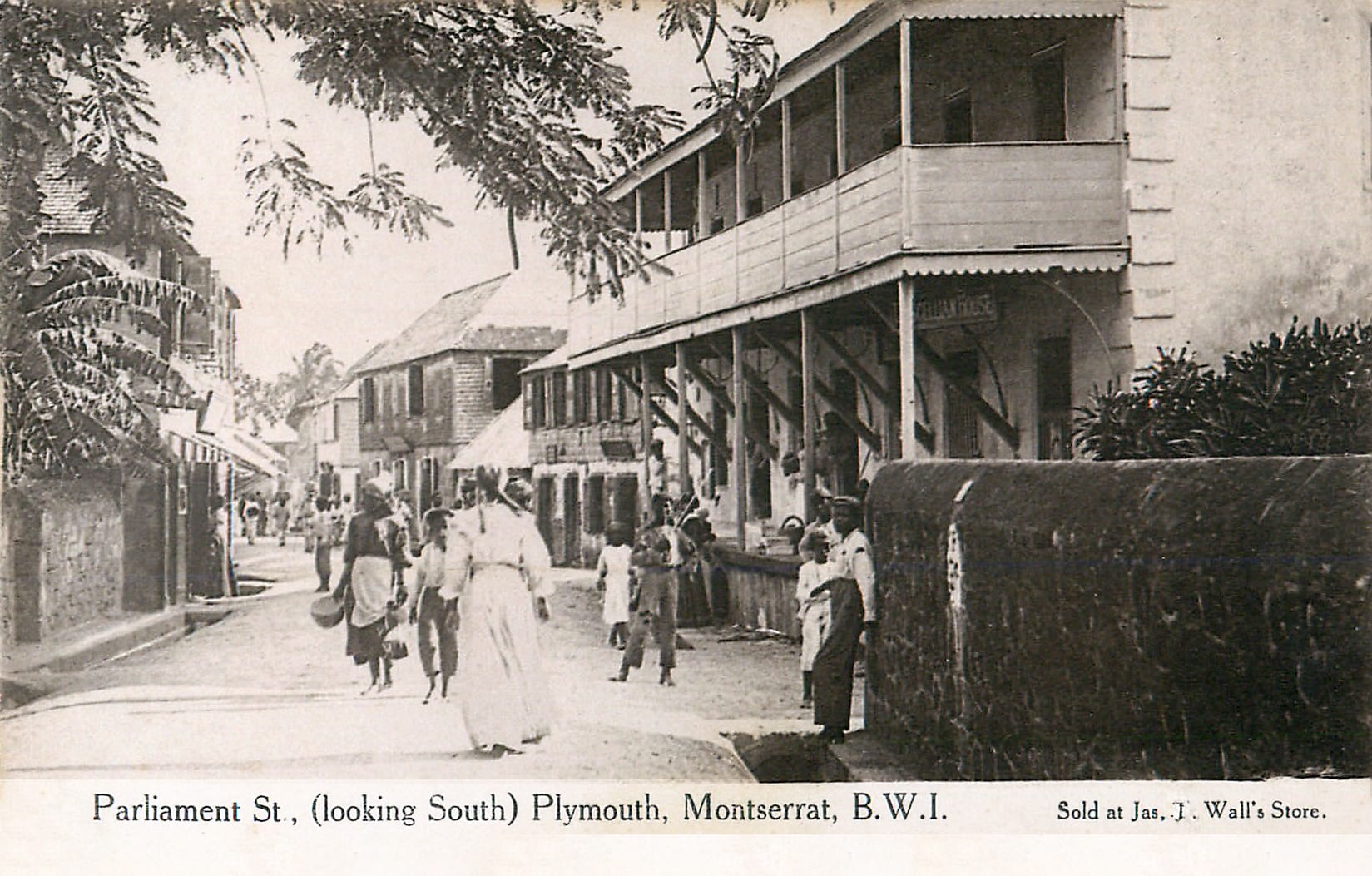 A black-and-white postcard of a street of two-storey buildings with wooden balconies, people in long dresses and hats walking along it under overhanging trees