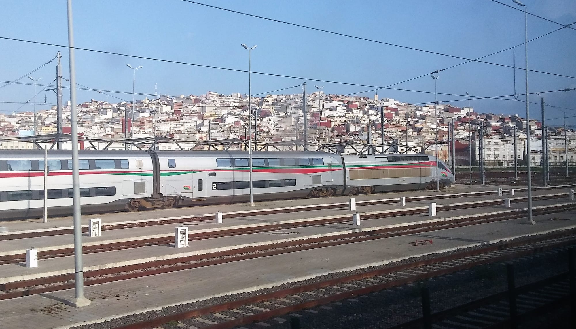 A modern high-speed train at a station platform in Tangier, with a dense hillside of white and red-roofed houses behind it