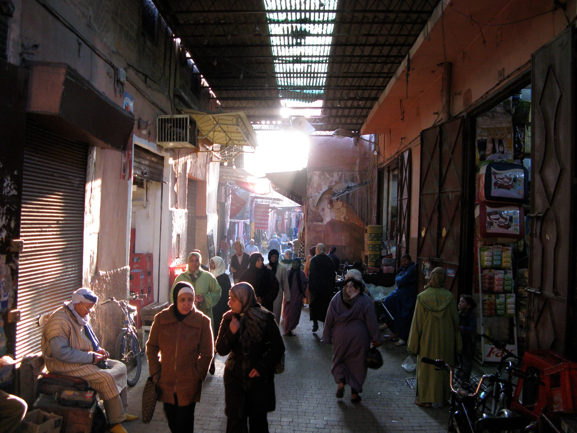 A covered souk alley crowded with people walking past shuttered and open shopfronts, with light filtering through a slatted roof