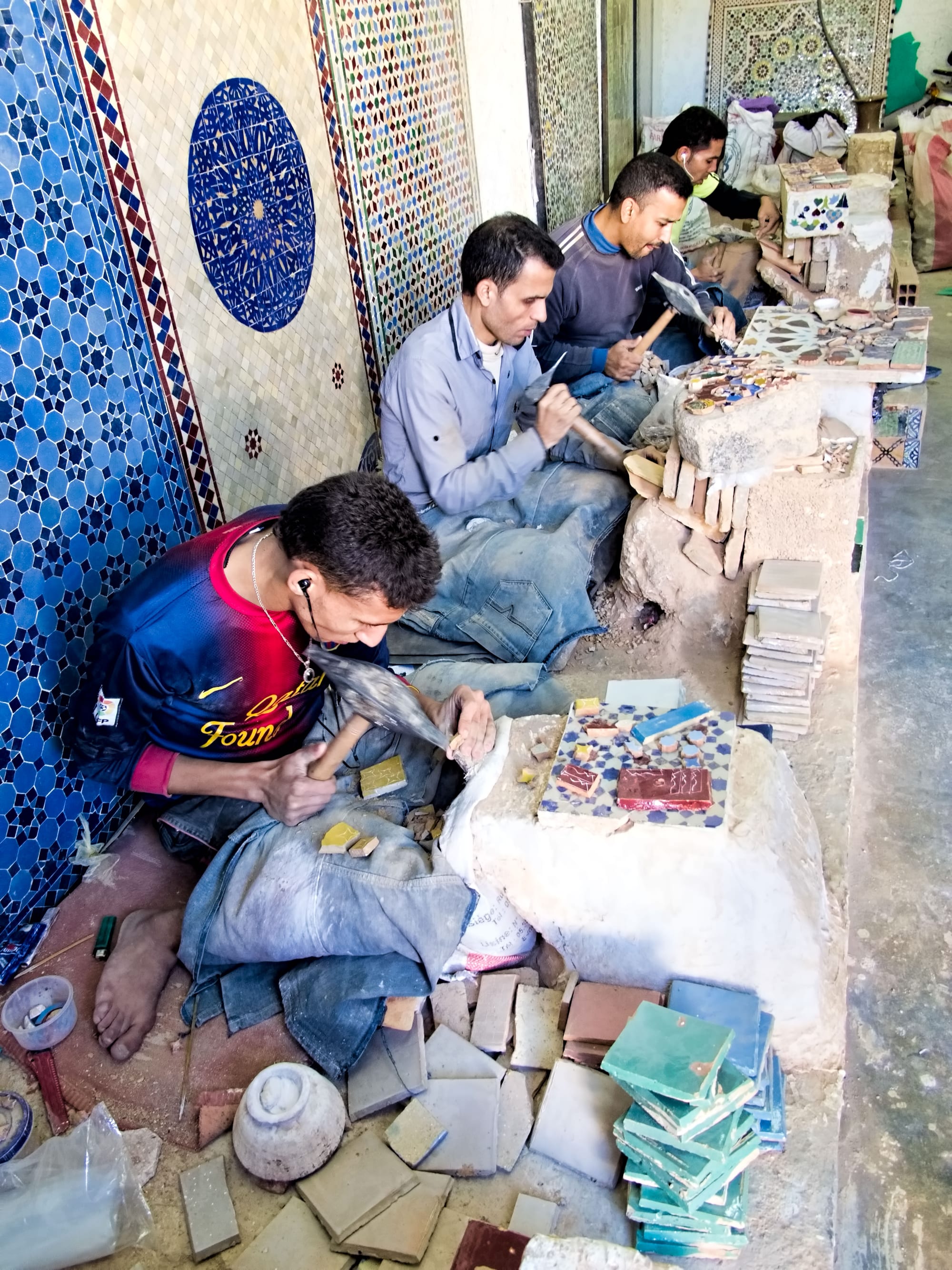 Three men sit on the floor of a workshop chipping glazed tiles into small shapes with hammers, with stacks of square green tiles in the foreground and finished blue and white mosaic panels on the wall beside them