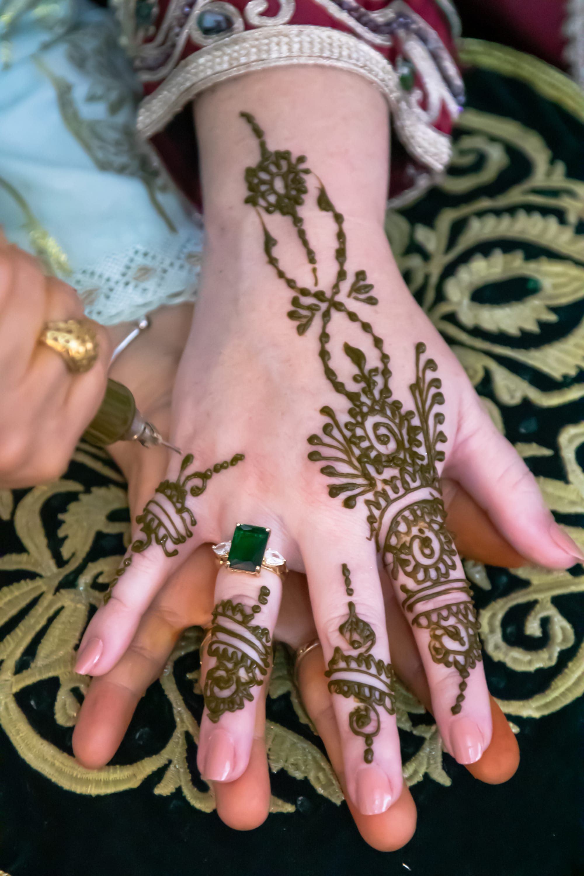 A bride's hand, wearing a green gemstone ring, rests on dark embroidered velvet while a woman's hand holds an applicator painting fine henna patterns across her fingers