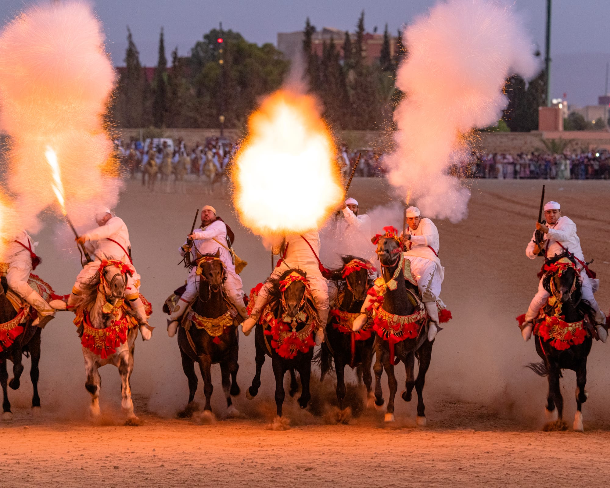 A line of riders in white robes and turbans gallop towards the camera on horses with red tasselled harnesses, their rifles firing bursts of flame and smoke, with a crowd of spectators behind