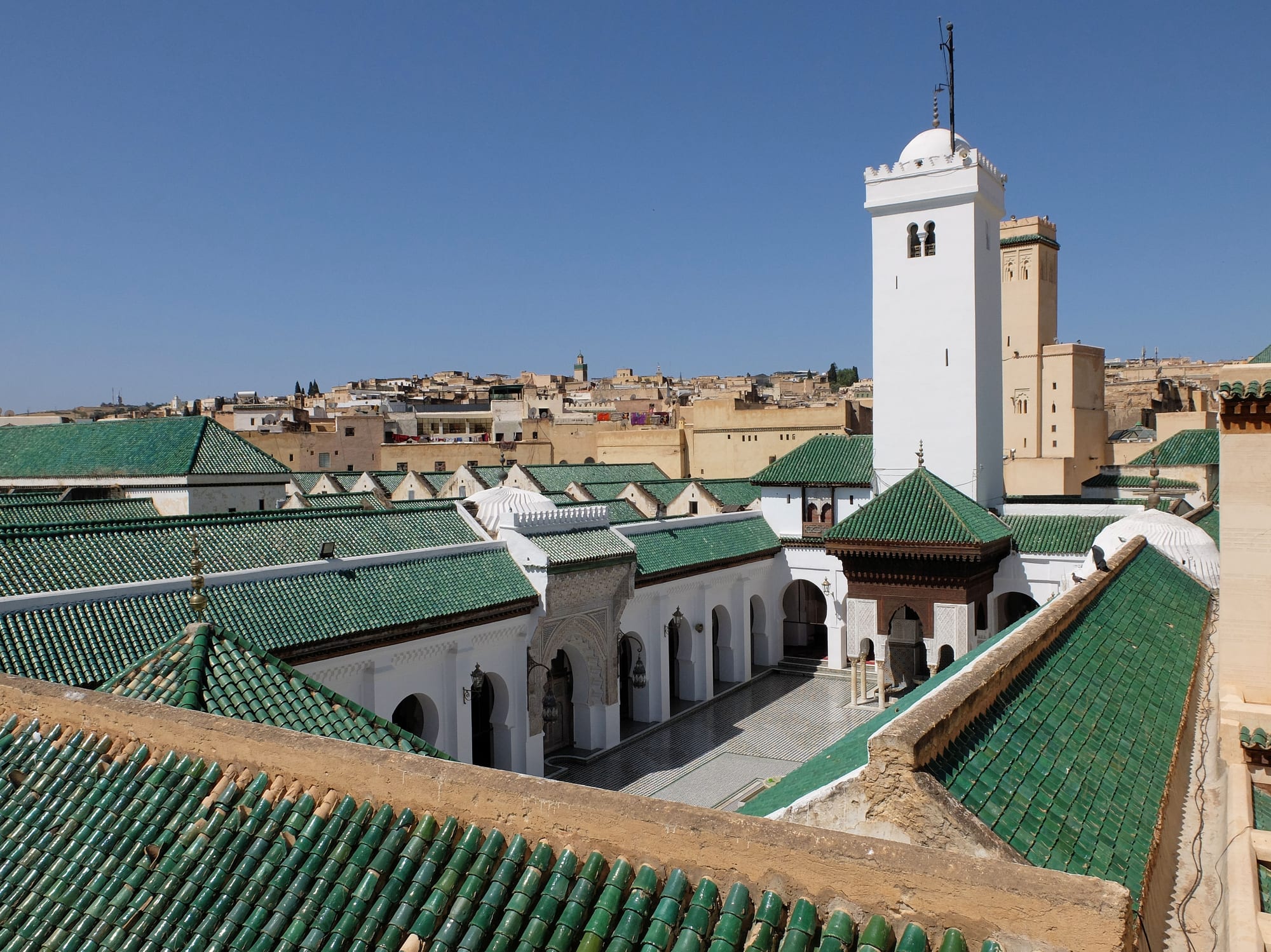 A rooftop view over green-tiled roofs into a mosque courtyard with white arcades and a carved fountain pavilion, with a tall white minaret and the old city spreading behind under a clear blue sky