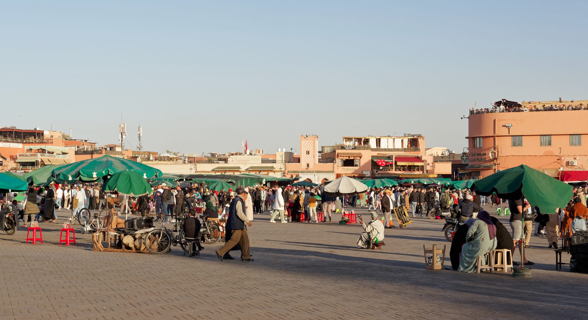 A huge open square packed with market stalls and crowds at dusk, mosque minaret behind