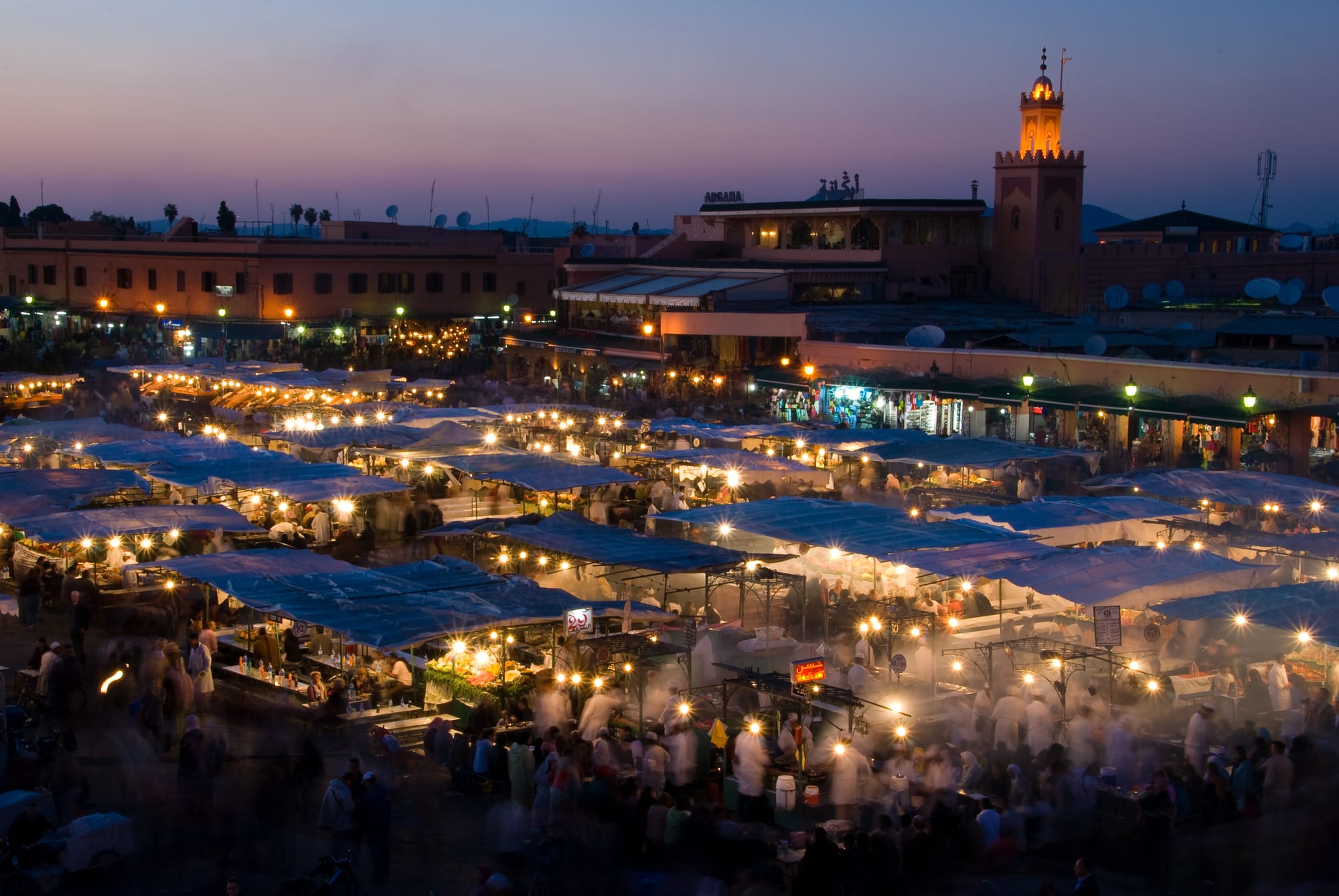 A vast square of lit food stalls and crowds at dusk, a minaret glowing behind