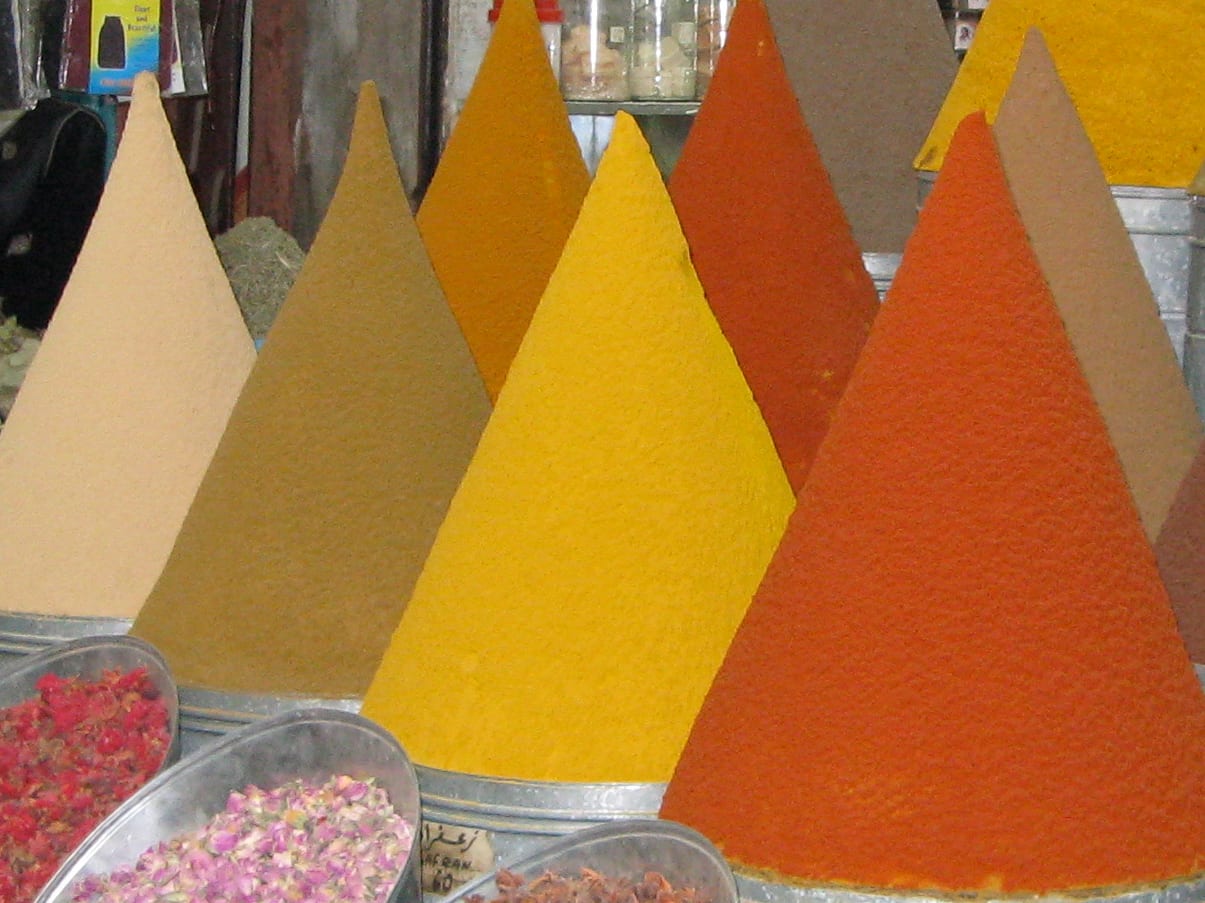Tall smooth cones of yellow, ochre and orange ground spice on a market stall, with metal trays of dried rosebuds and red threads in front