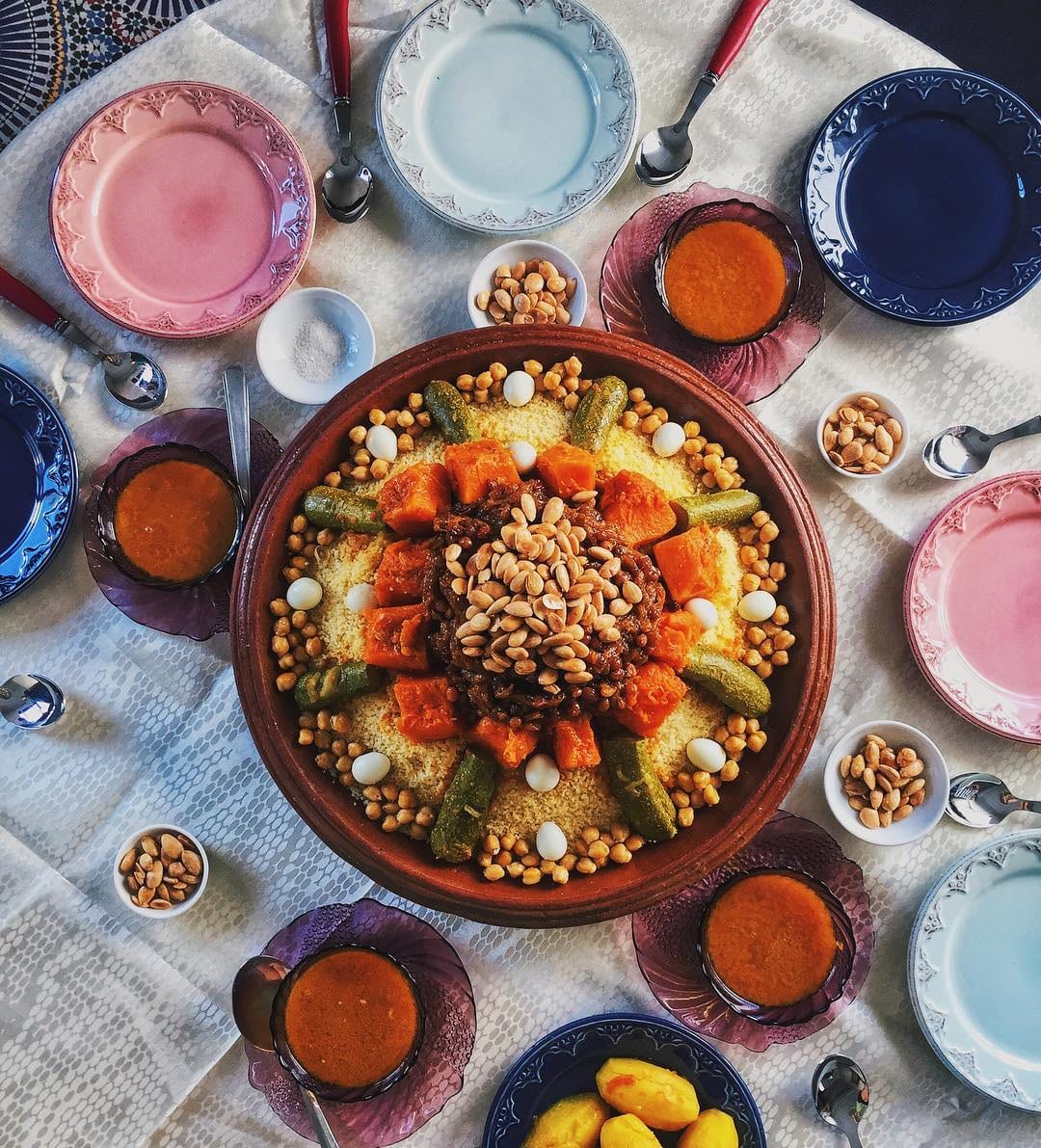 An overhead view of a large round platter of couscous topped with squash, courgette, okra, chickpeas and a dark onion topping, ringed by spoons and empty plates