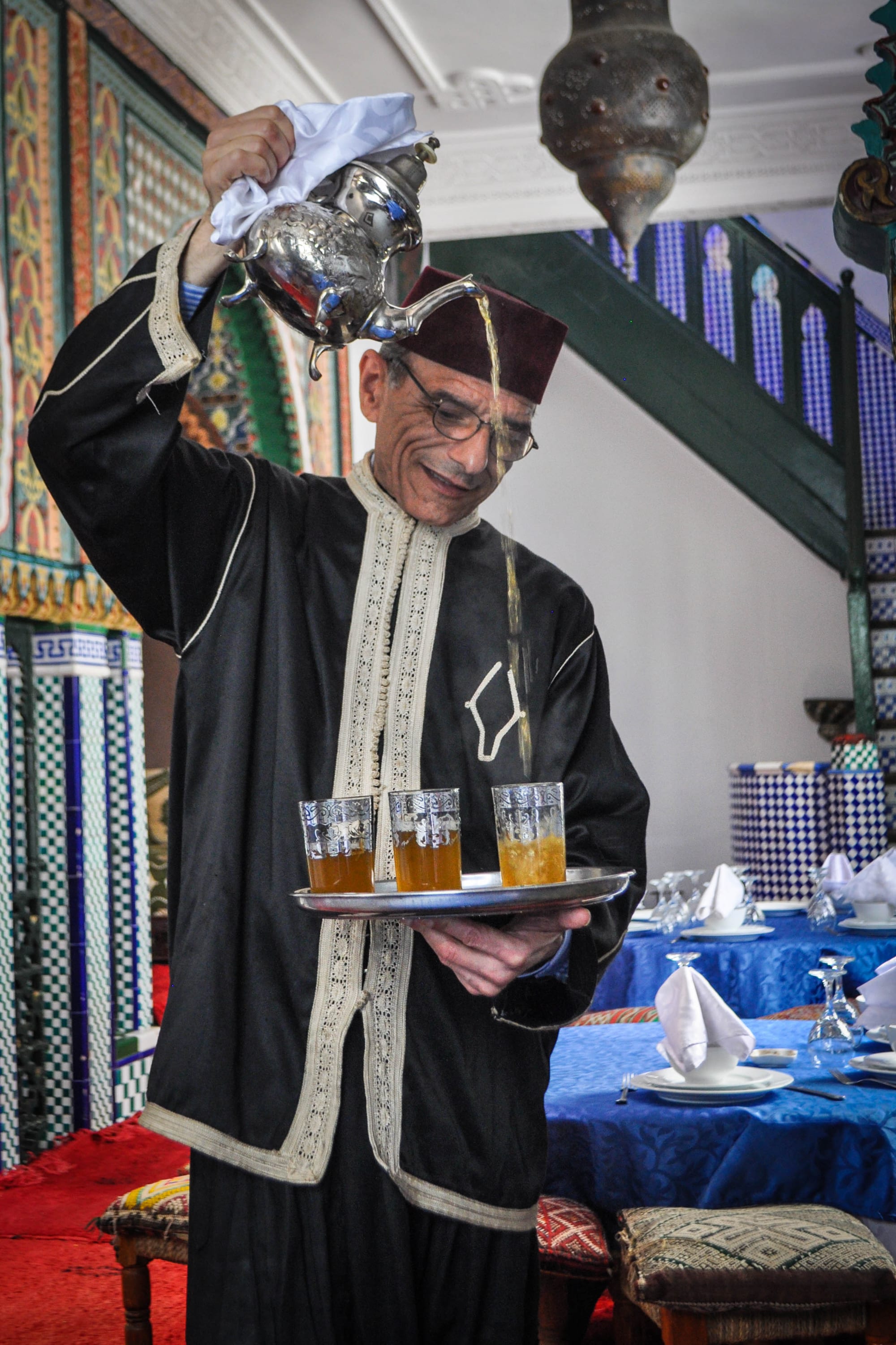 A man in a dark robe and burgundy fez pouring tea in a thin stream from a raised silver pot into small glasses on a tray