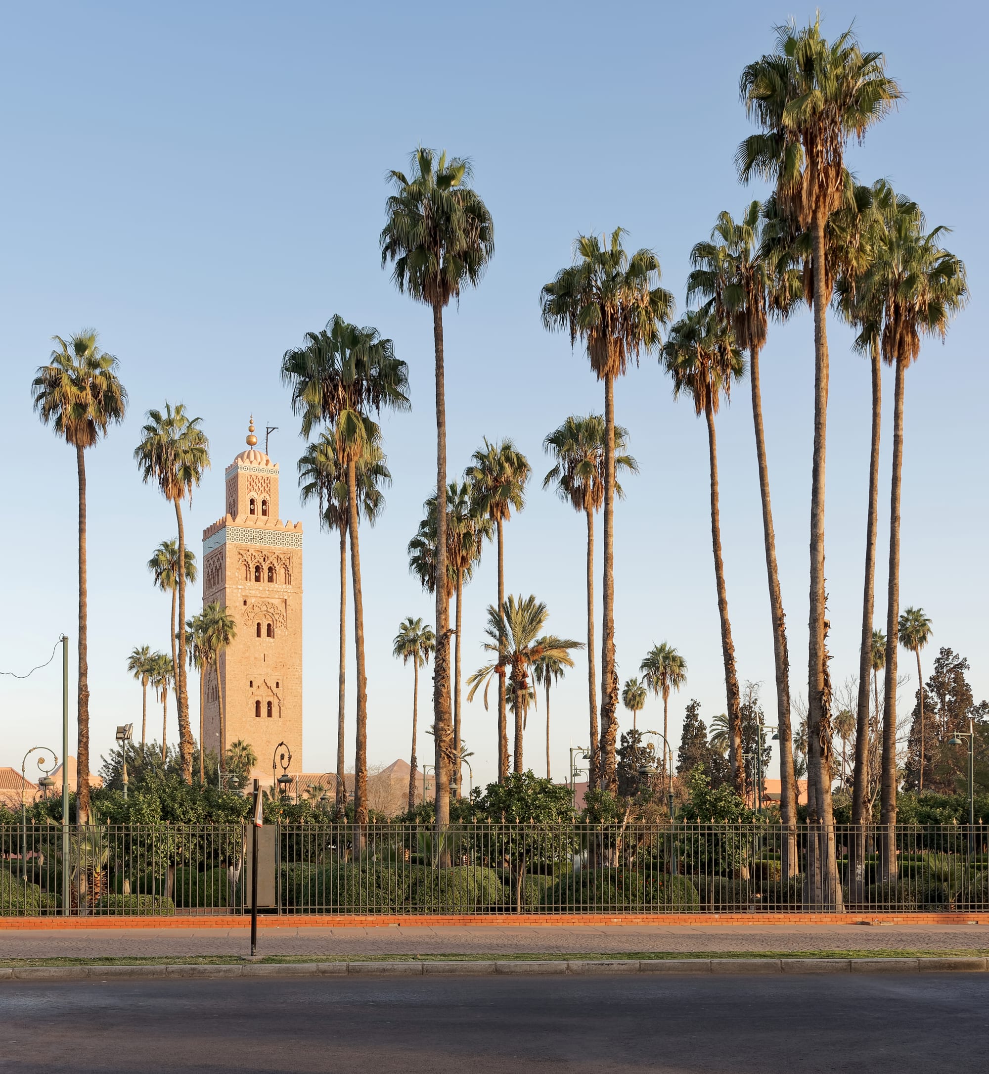 A tall sandstone minaret with blind arches and a green-tiled band, rising above palm trees