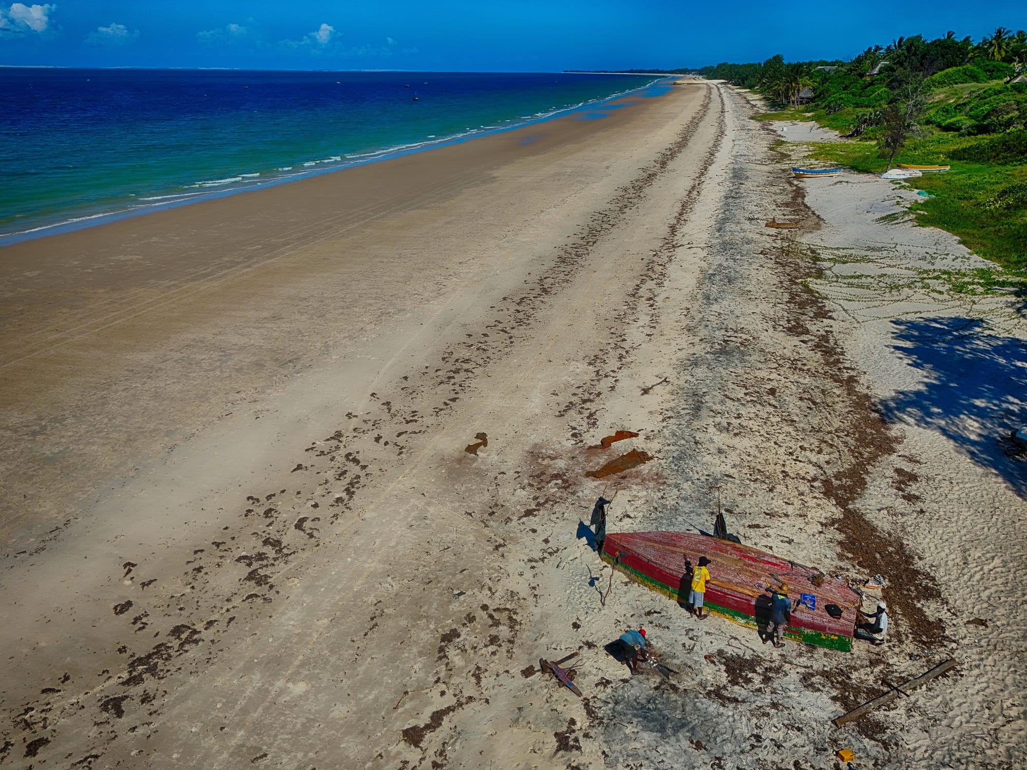Aerial view of fishermen mending a red and green wooden fishing boat on a wide white sand beach beside turquoise water