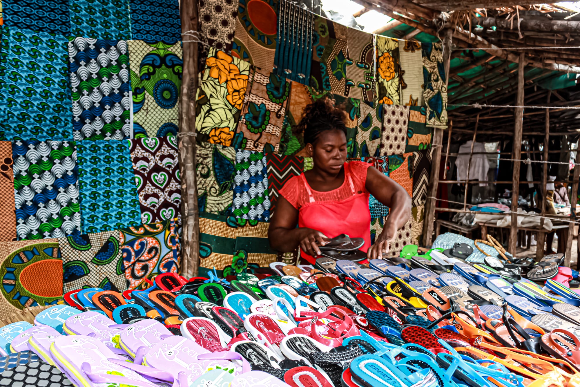 A market vendor arranging colourful printed capulana cloth and rows of sandals at an open-air stall
