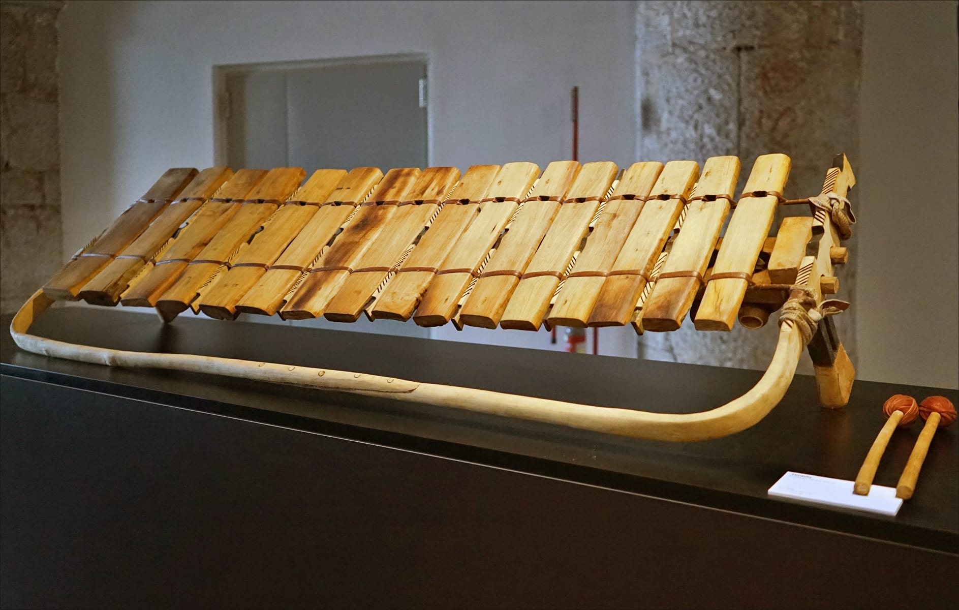 A wooden xylophone with a row of pale wooden keys lashed to a long curved frame, with two beaters resting beside it on a black plinth