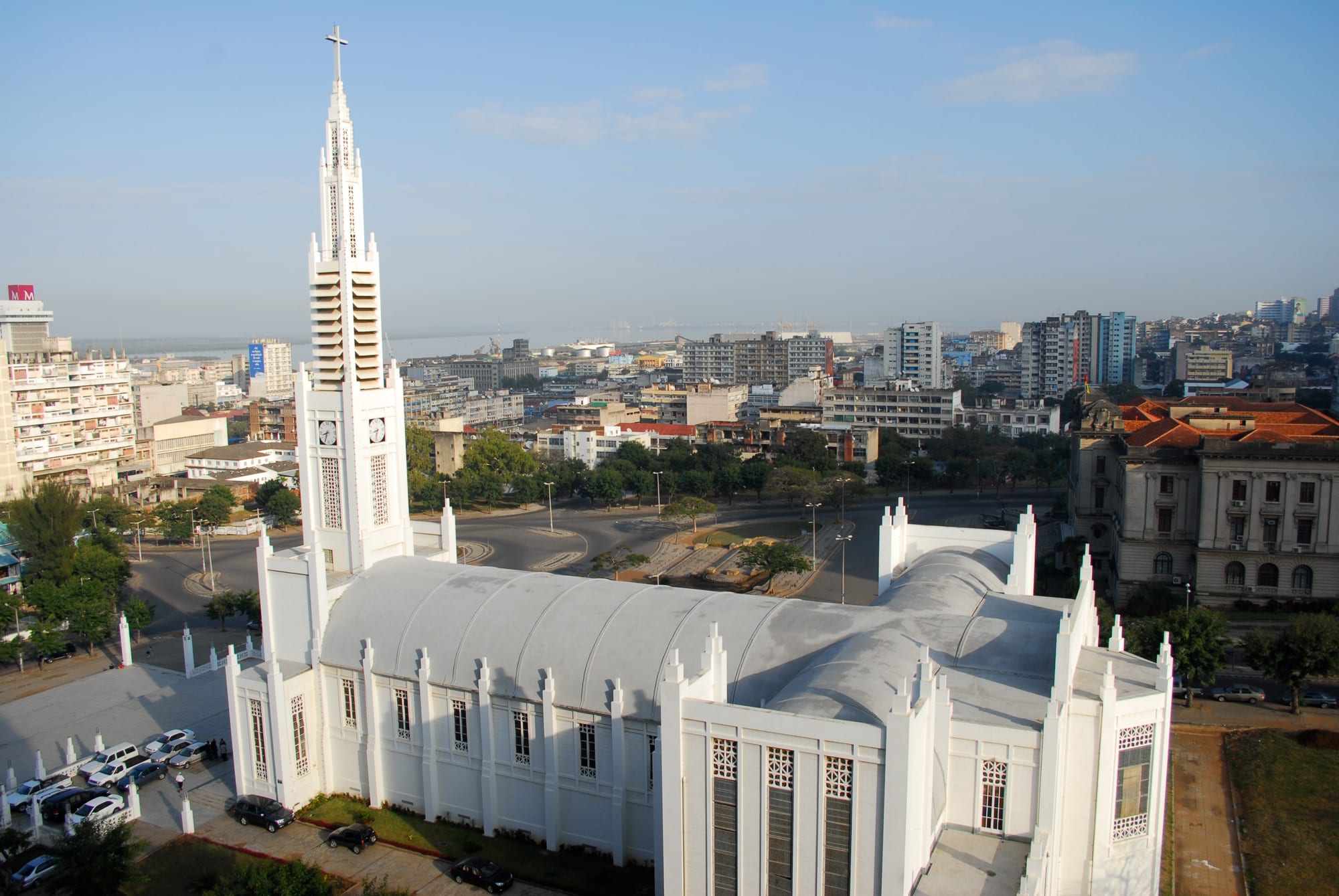 A large white church with a tall, slender spire topped by a cross, seen from above, with city blocks and a hazy bay stretching behind it