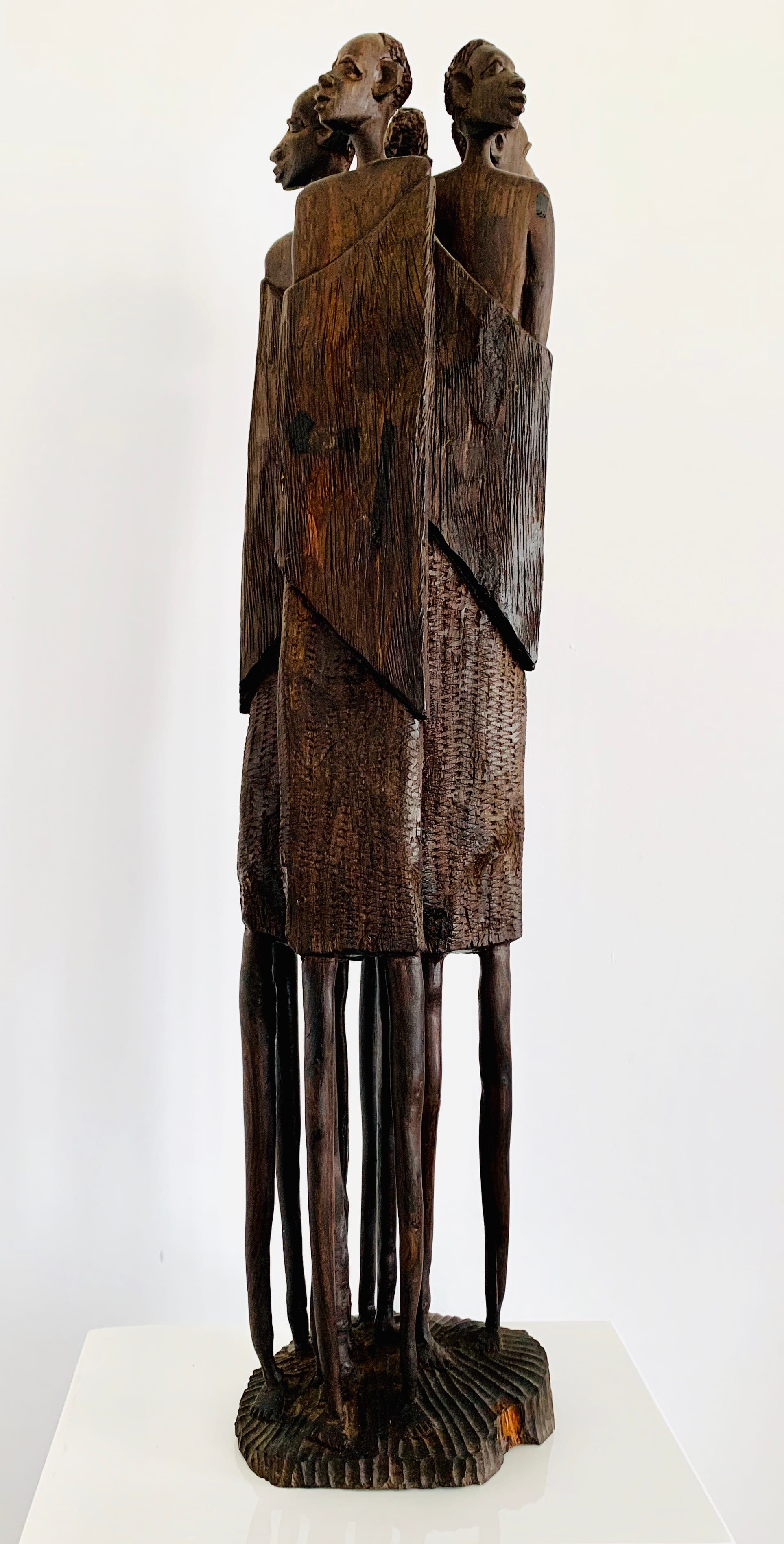 A tall, dark wooden carving of several slender figures pressed together shoulder to shoulder, their long thin legs rising from a single carved base