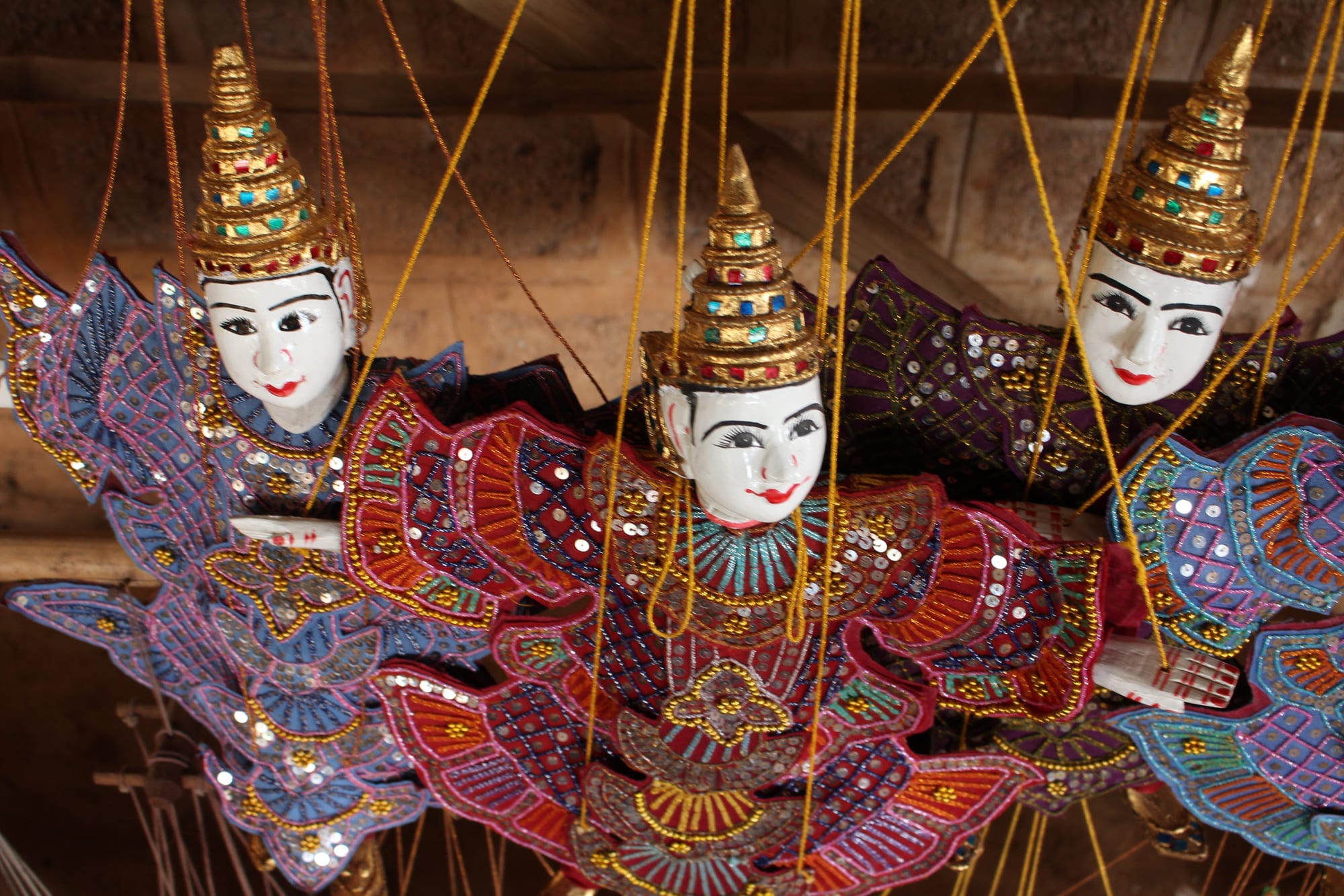 Three string puppets hang side by side, with white painted faces, red lips, tall gold jewelled crowns and richly sequined costumes in red, purple and blue, their golden strings rising out of the frame