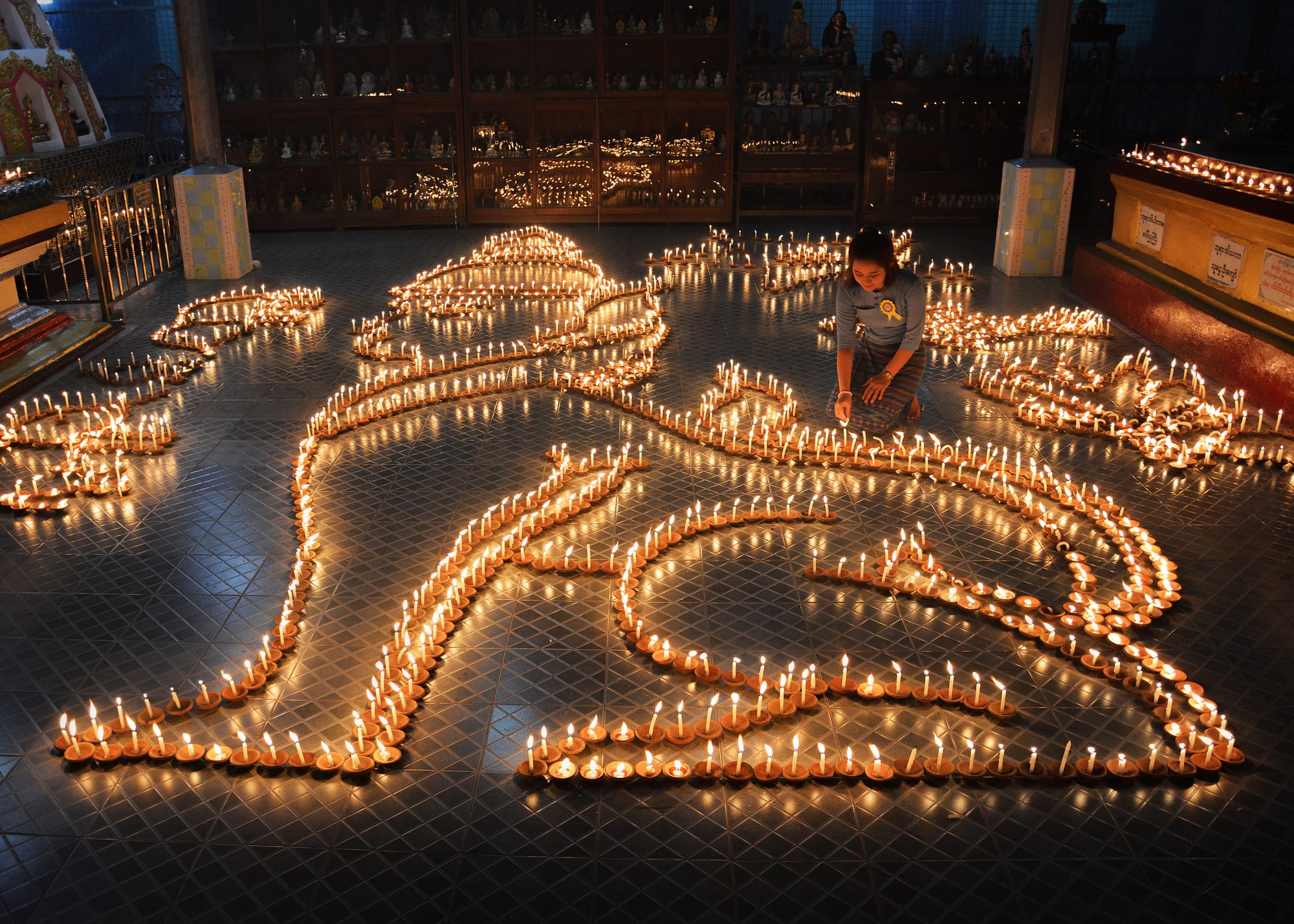 A woman kneels on a tiled floor lighting hundreds of small oil lamps laid out in the shape of a large lotus and other flowing patterns, with shelves of small Buddha images behind her