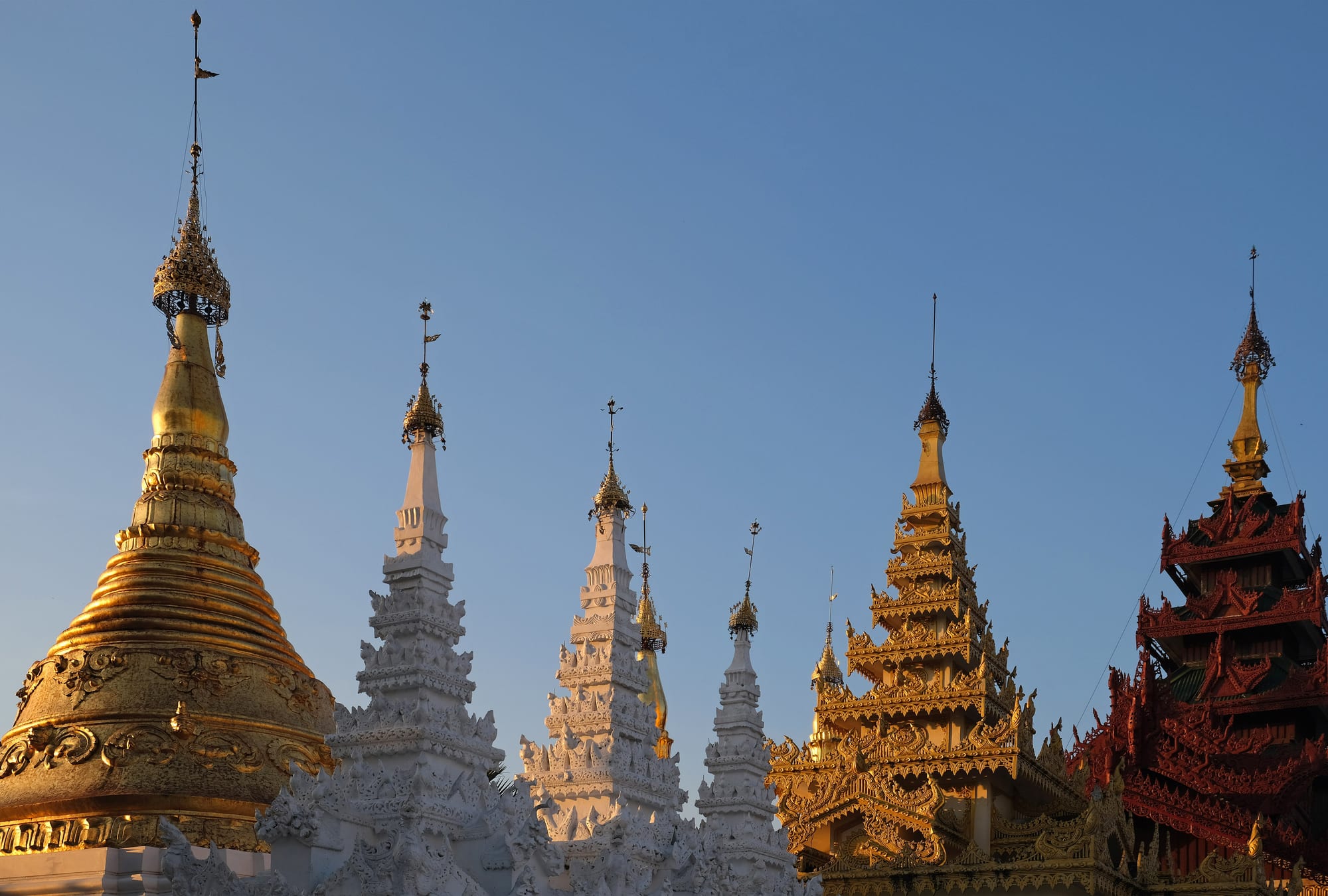 A row of pagoda spires against a clear evening sky — a gilded bell-shaped stupa on the left, three white carved spires in the middle and tiered gold and red roofs on the right