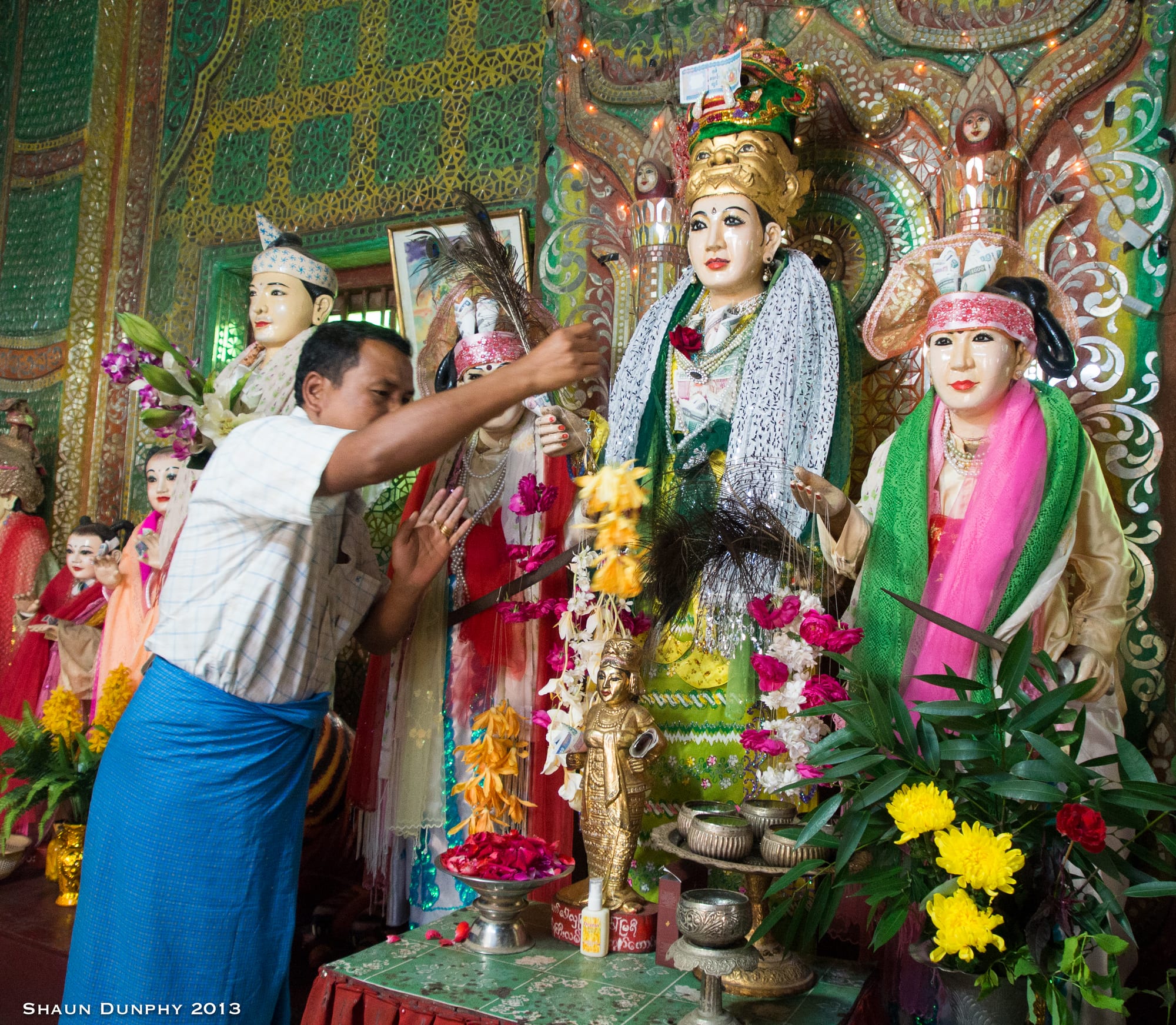A man in a blue longyi places flowers among brightly painted, life-size statues of spirit figures in crowns and silk robes, in a shrine decorated with mirrored mosaic