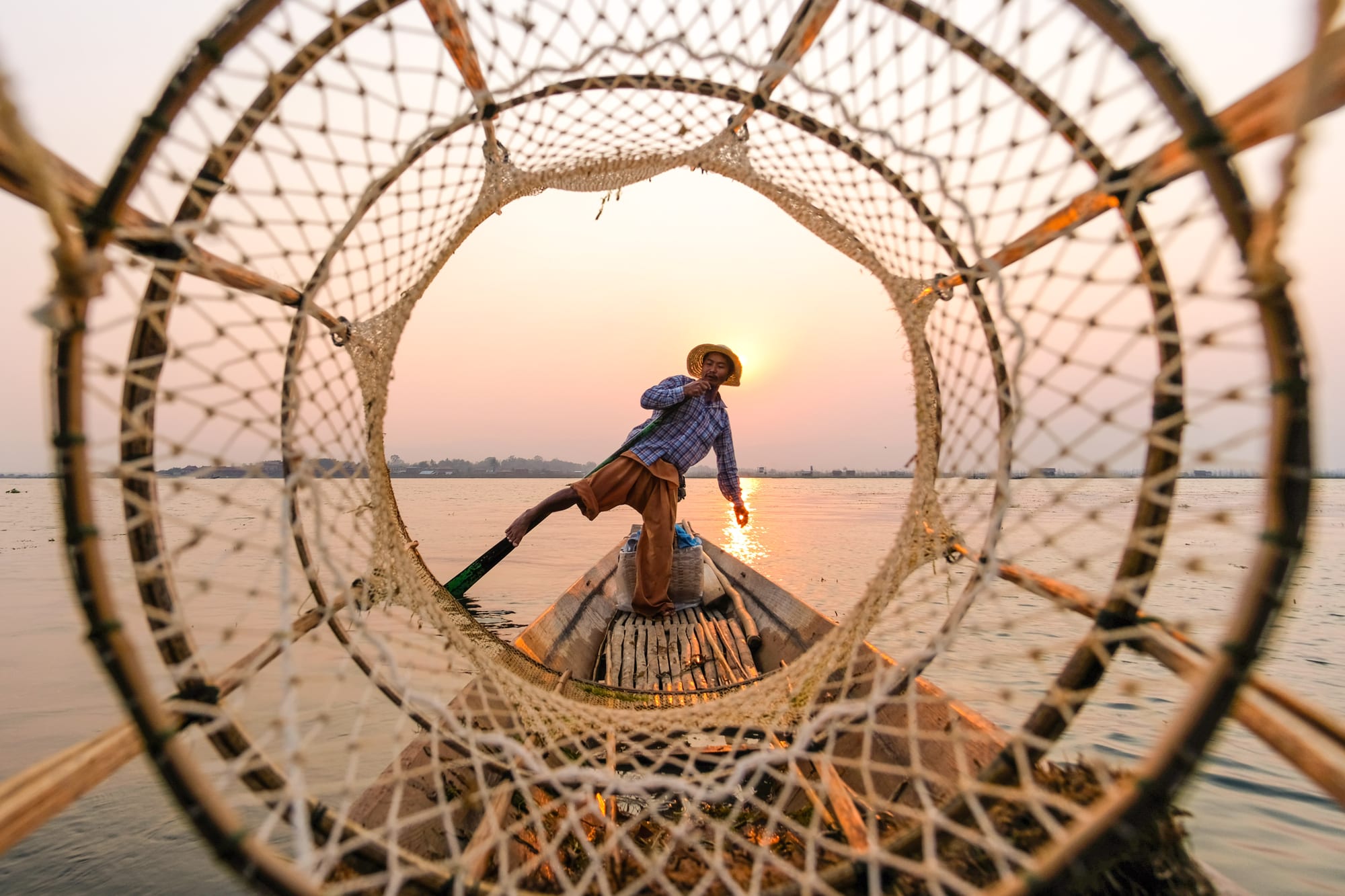 A fisherman balancing on one leg while rowing a boat on a calm lake