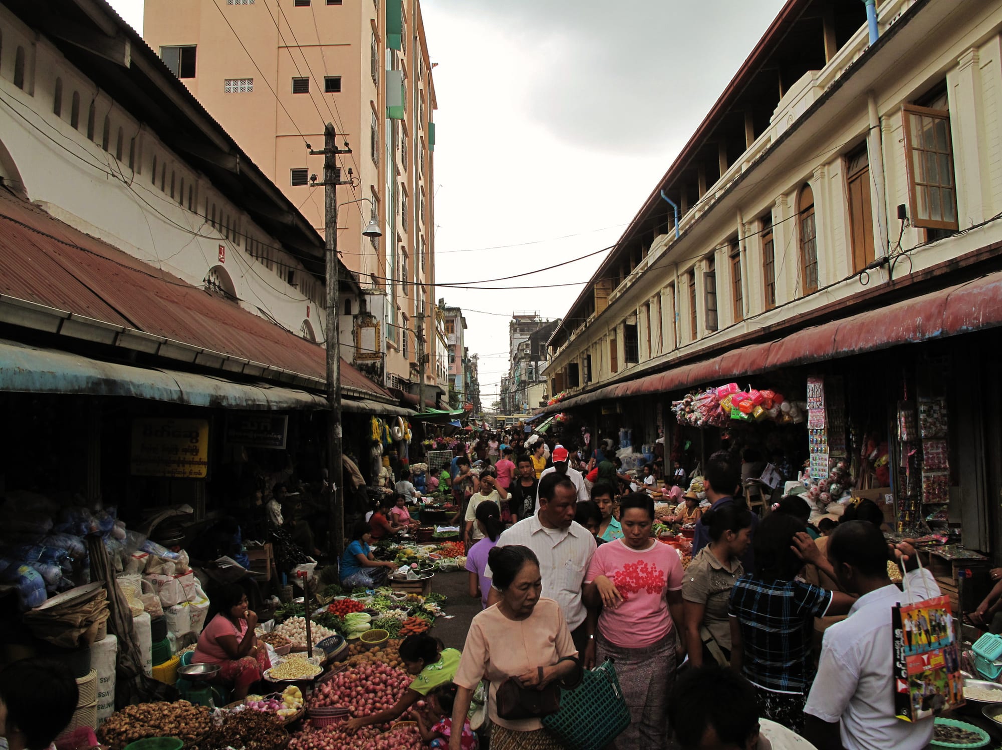 A crowded narrow market street in Yangon lined with covered stalls of vegetables and goods, shoppers and vendors filling the lane