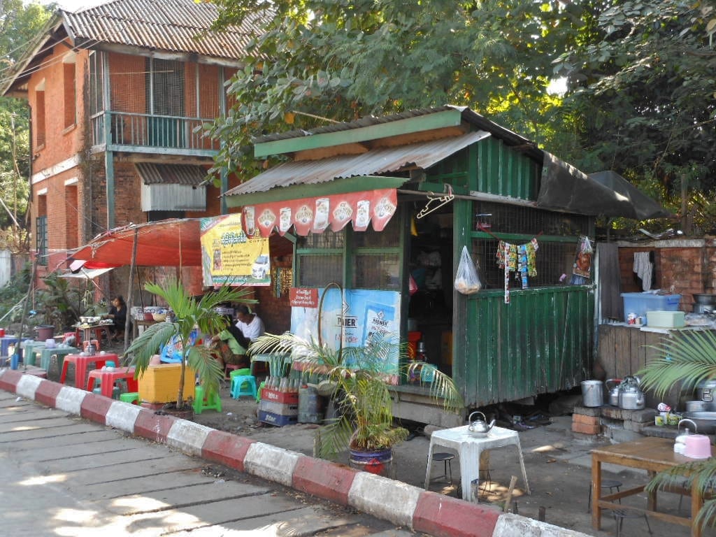 A roadside teahouse stall with a green corrugated awning, low plastic stools and tables, and kettles set out for tea