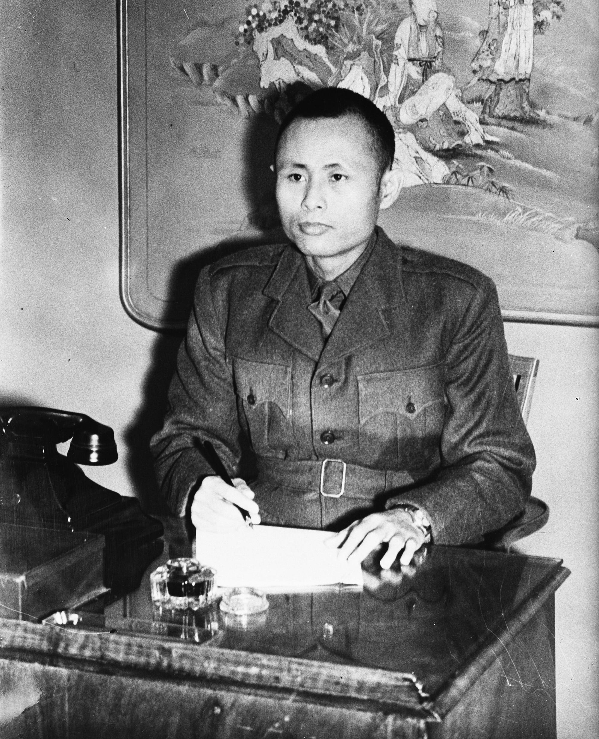 A black-and-white photograph of a young man in a belted military tunic seated at a desk, pen in hand over a sheet of paper, a telephone beside him
