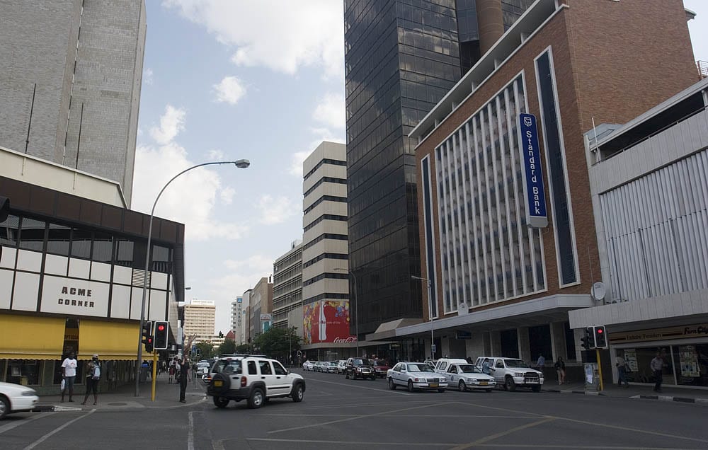 A city street in downtown Windhoek lined with mid-rise office buildings, cars waiting at a junction and pedestrians on the pavement