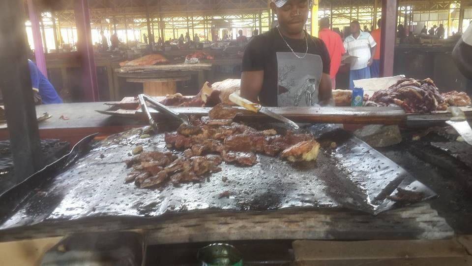 A vendor grilling beef on a large open griddle at an outdoor kapana stall in Katutura, with other stalls and customers behind him
